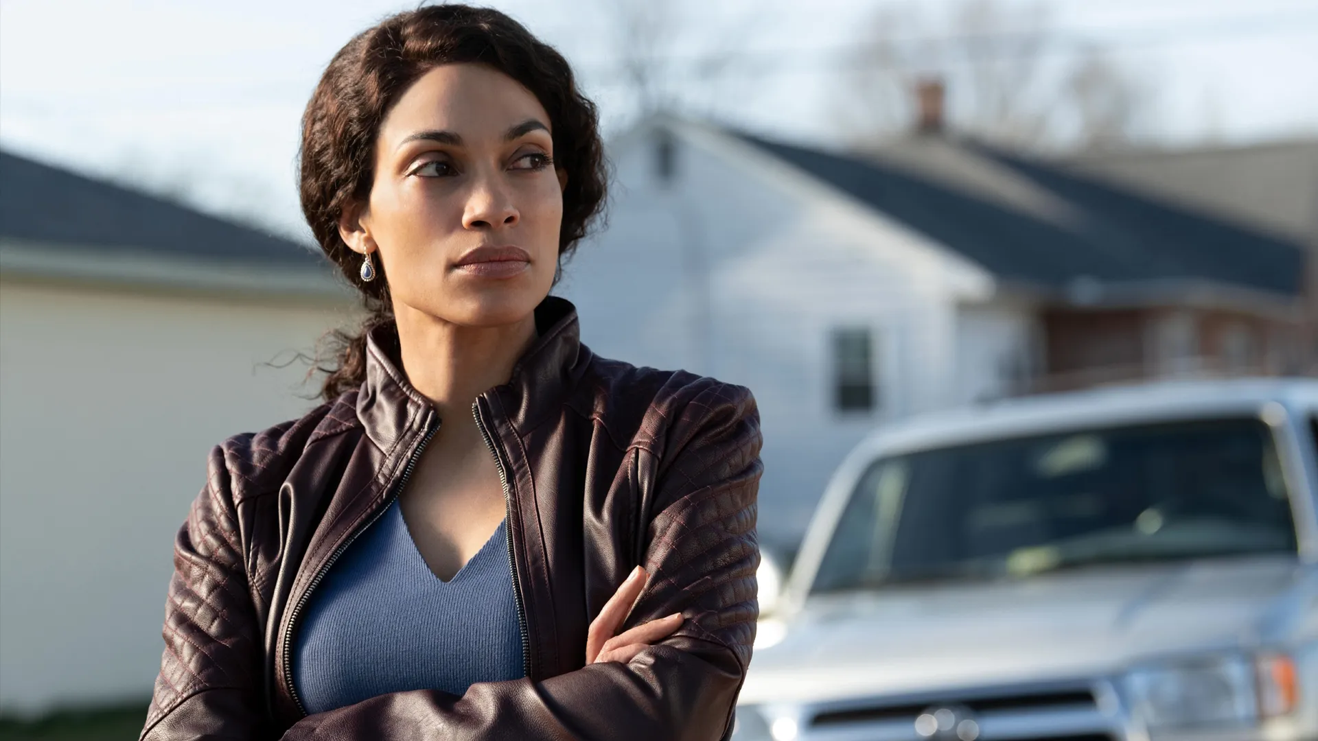 A thoughtful woman, Bridget Meyer (Rosario Dawson), stands outdoors with her arms crossed while wearing a dark brown leather jacket and looking off to the side with suburban houses and a silver vehicle blurred in the background.