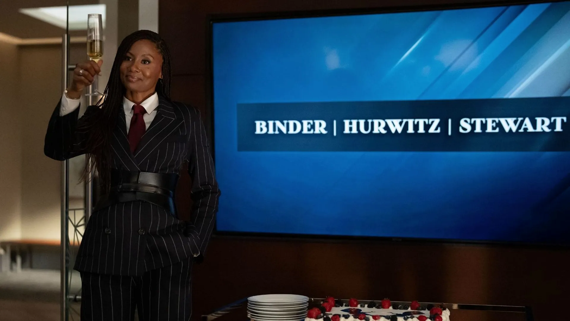 Jax Stewart, segurando uma taça de champanhe diante de uma tela que exibe o nome do escritório: Binder | Hurwitz | Stewart.