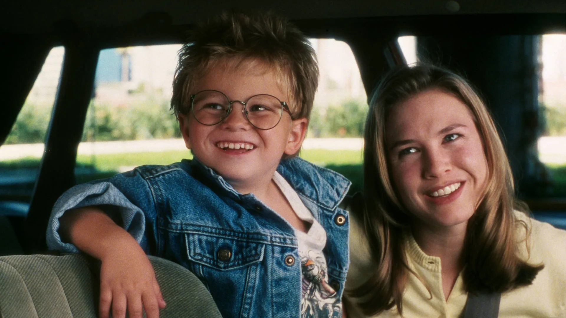 A young Jonathan Lipnicki with spiky blonde hair and large round glasses sits next to a smiling Renée Zellweger inside a car from the 1996 film Jerry Maguire.