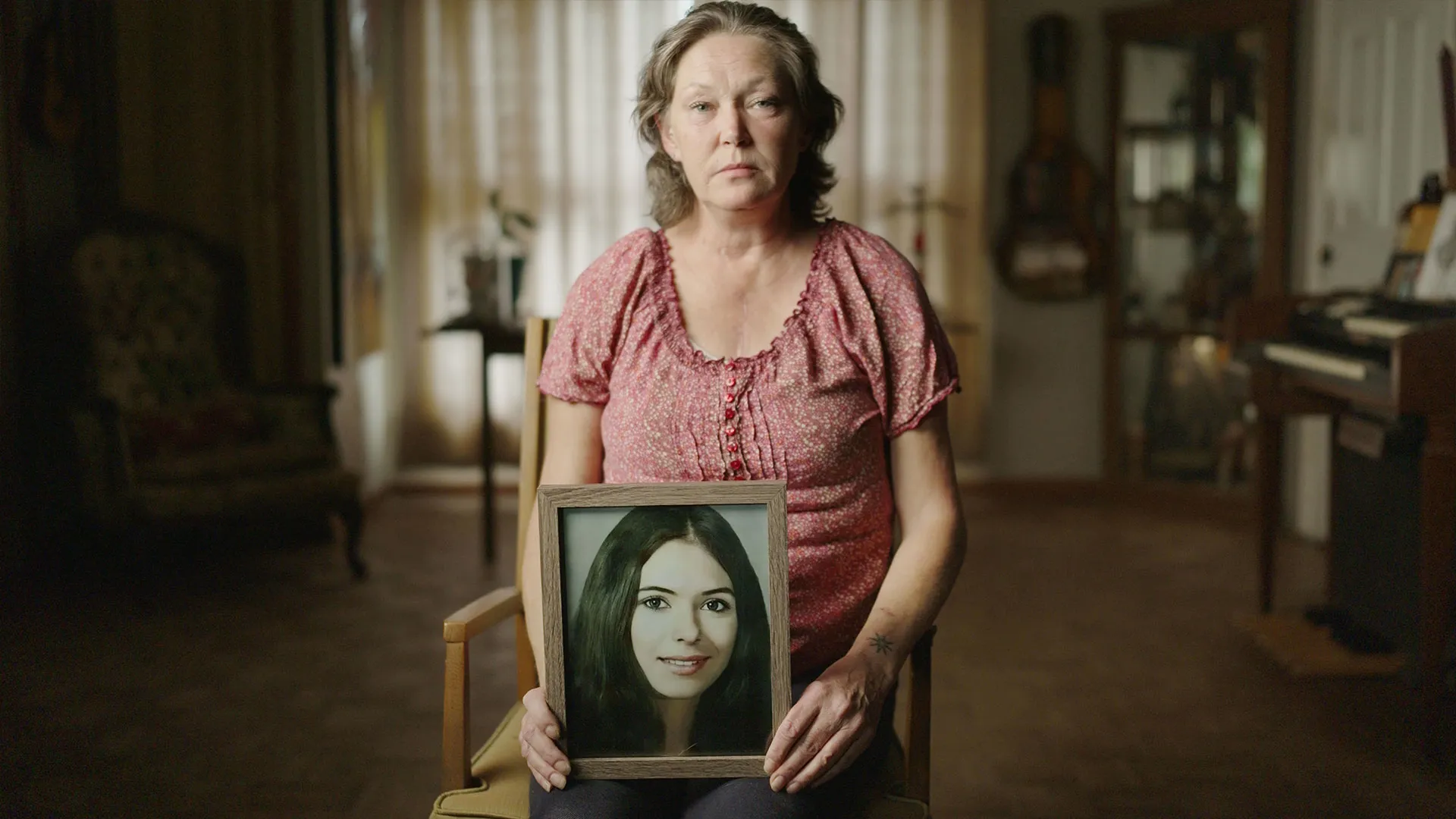 Key art for Naming The Dead featuring a seated woman holding a framed portrait photograph in a living room.