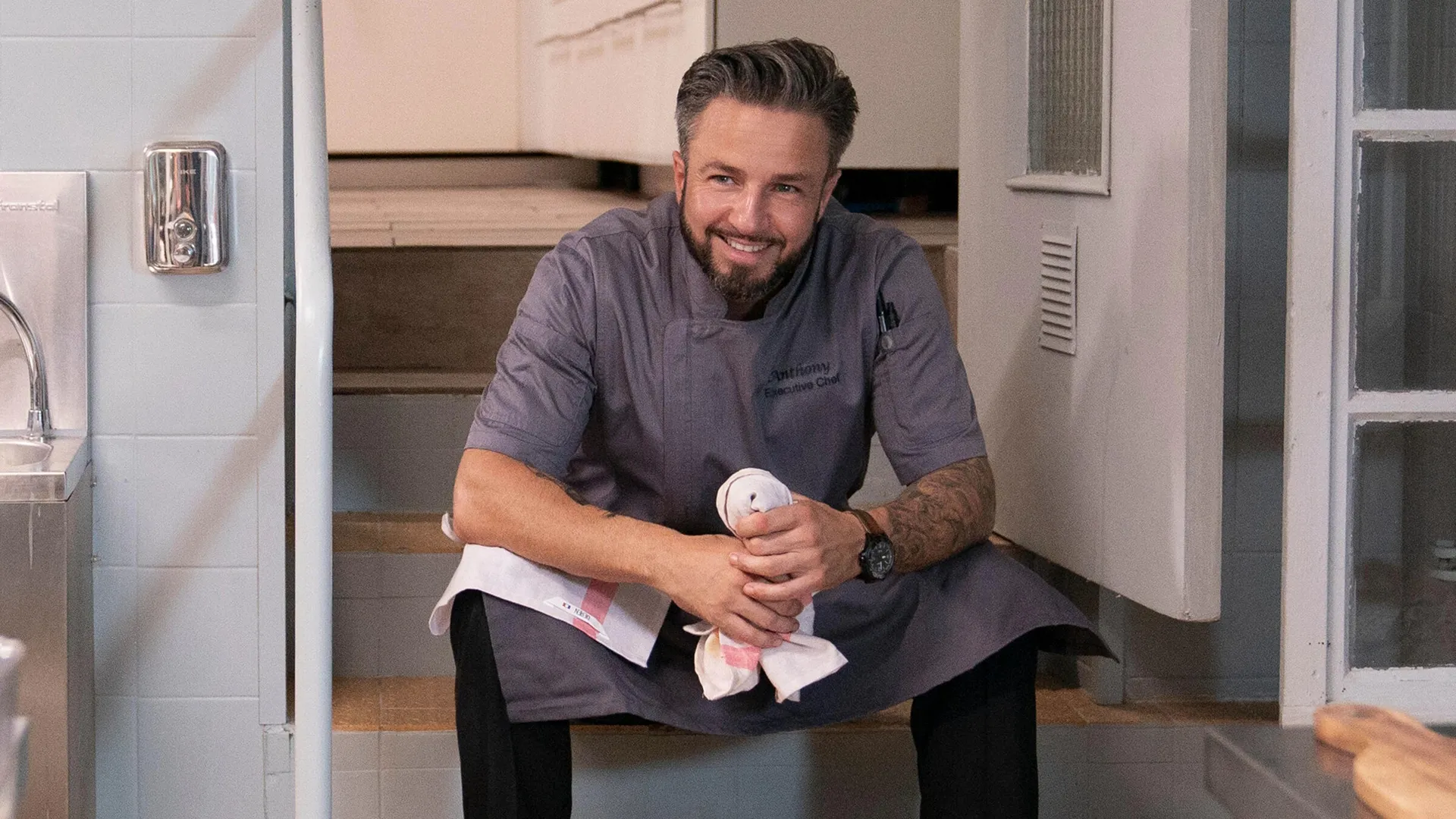 Anthony smiles sitting on the steps of the kitchen in his chef uniform