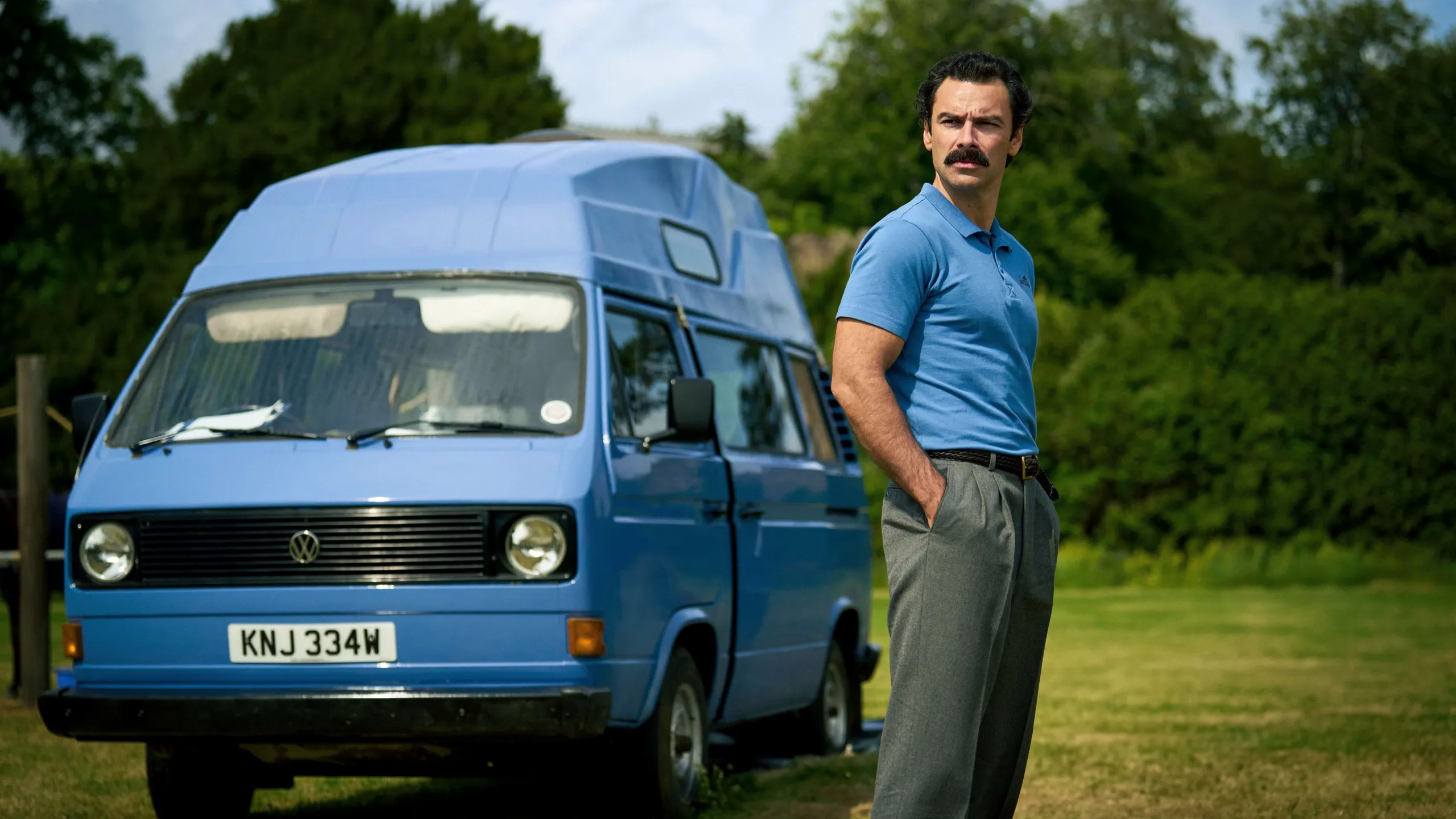 Declan O’Hara (Aidan Turner) stands beside a blue Volkswagen camper van outdoors in Rivals season 2.
