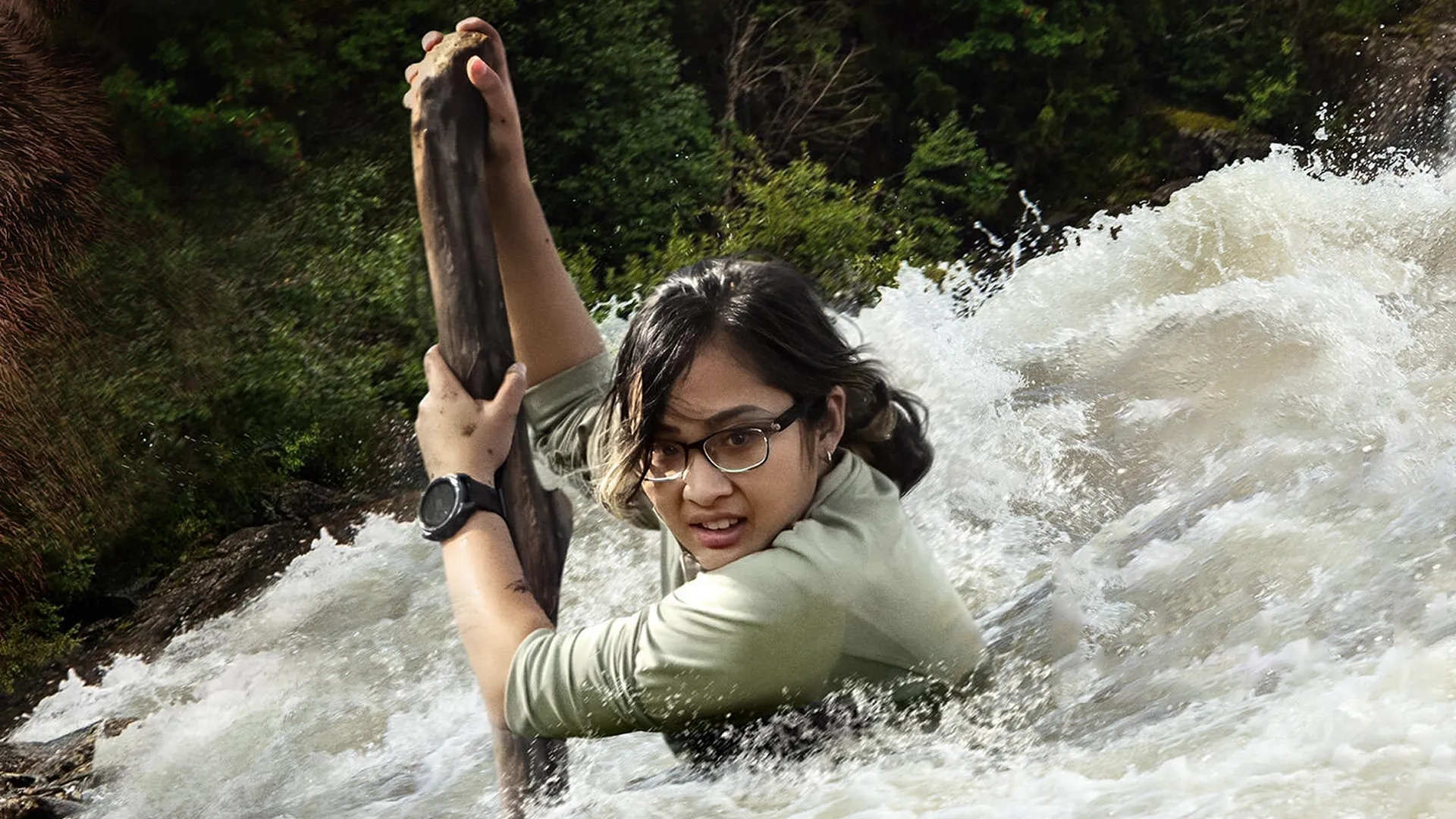 A woman clings to a stick while in a river rapids with a determined look on her face.