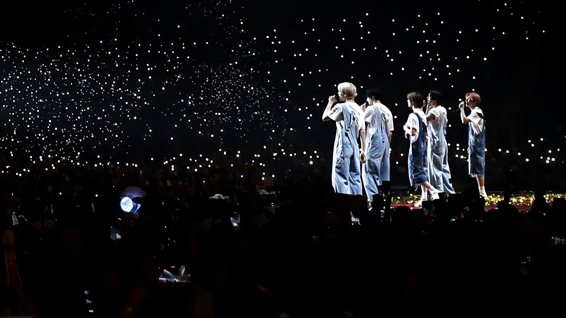 The group perform on stage in a dark arena illuminated by lights from the crowd.
