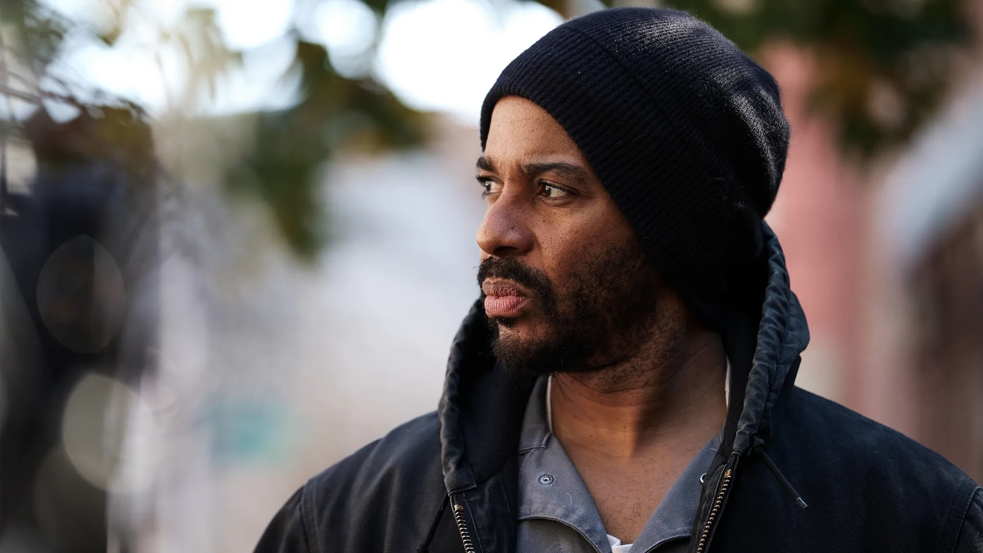 A close-up profile shot of a Black man with a beard, wearing a black knit beanie and a dark, heavy jacket over a collared shirt. 