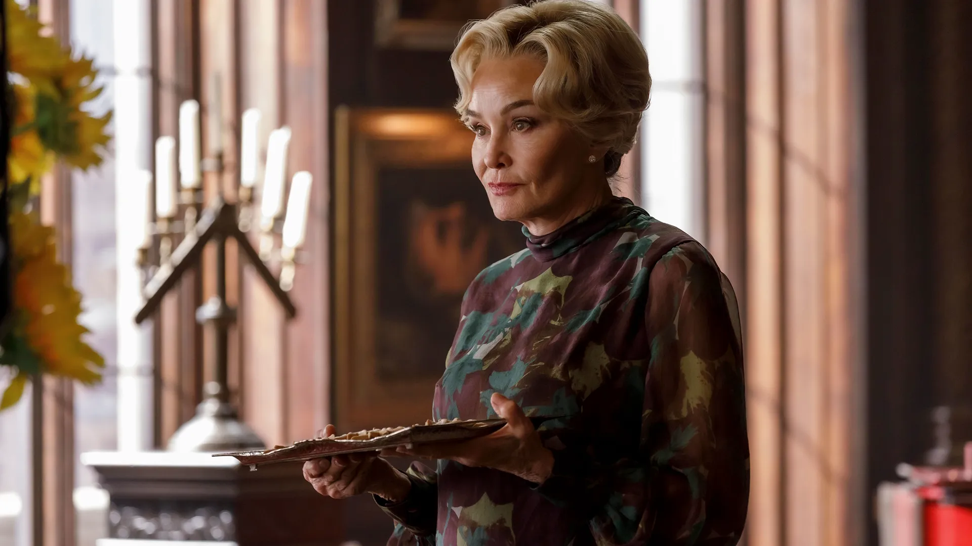 A mature blonde woman with styled, coiffed hair looks off to the side while holding a flat serving tray, wearing a high-necked, long-sleeved sheer blouse with an abstract floral pattern in shades of green and maroon.