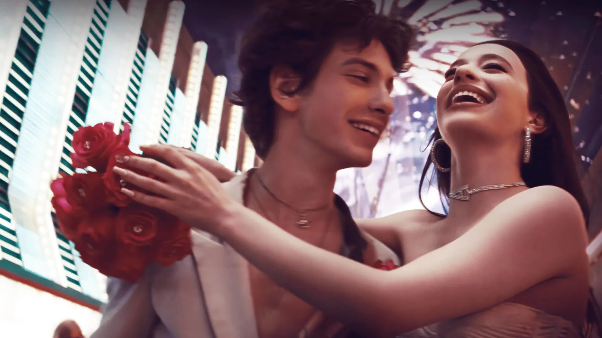 Mikey Madison as Anora and Mark Eydelshteyn as Ivan smiling and embracing with red roses against a blurred Las Vegas neon background in Sean Baker's movie Anora.