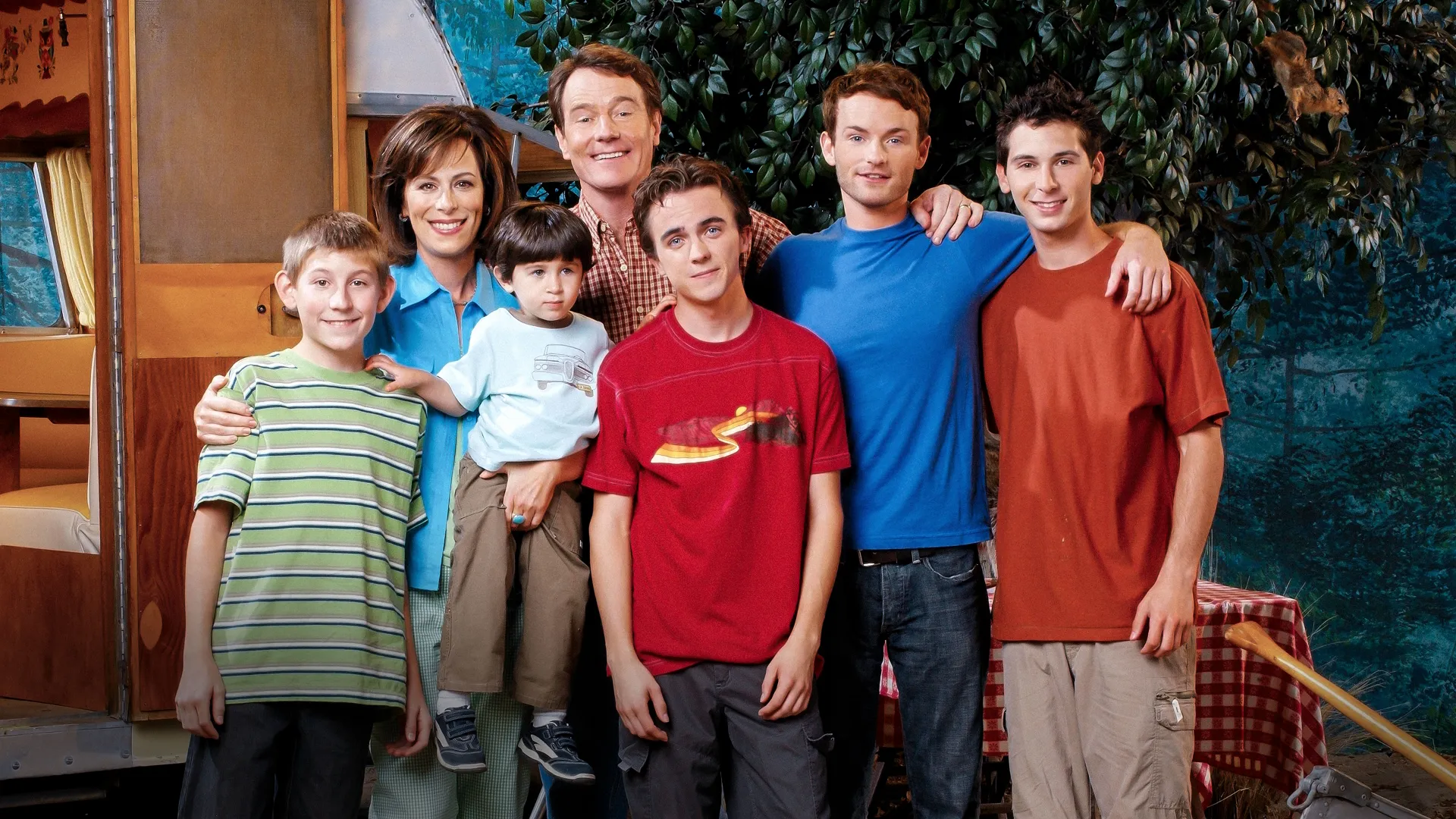 Una foto promocional del elenco de la sitcom Malcolm in the Middle muestra a Hal (Bryan Cranston) y Lois (Jane Kaczmarek) de pie con sus cinco hijos: Dewey (Erik Per Sullivan), Jamie (James y Lukas Rodriguez), Malcolm (Frankie Muniz), Francis (Christopher Masterson) y Reese (Justin Berfield), sonriendo juntos frente a una caravana al aire libre y una zona de pícnic