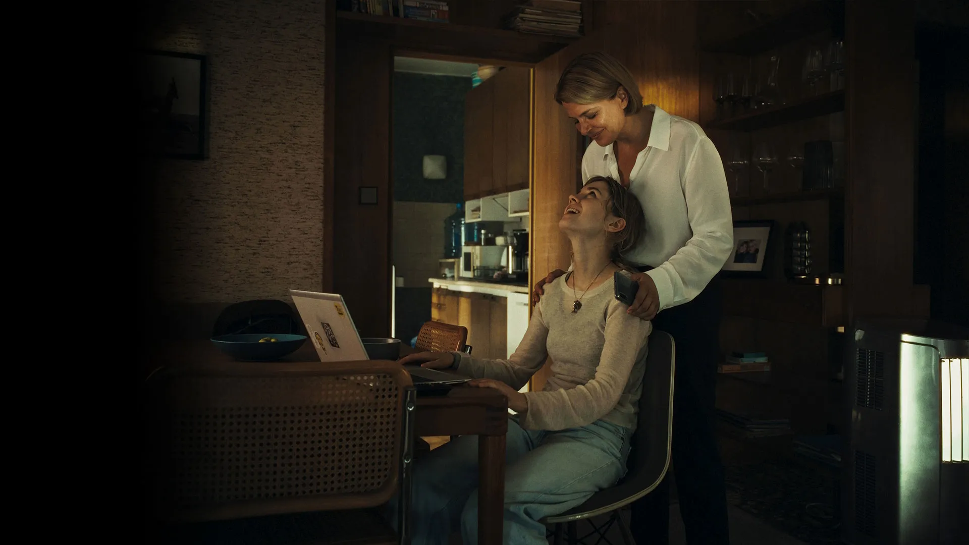 One person stands beside another seated at a dining table using a laptop in a dimly lit home.
