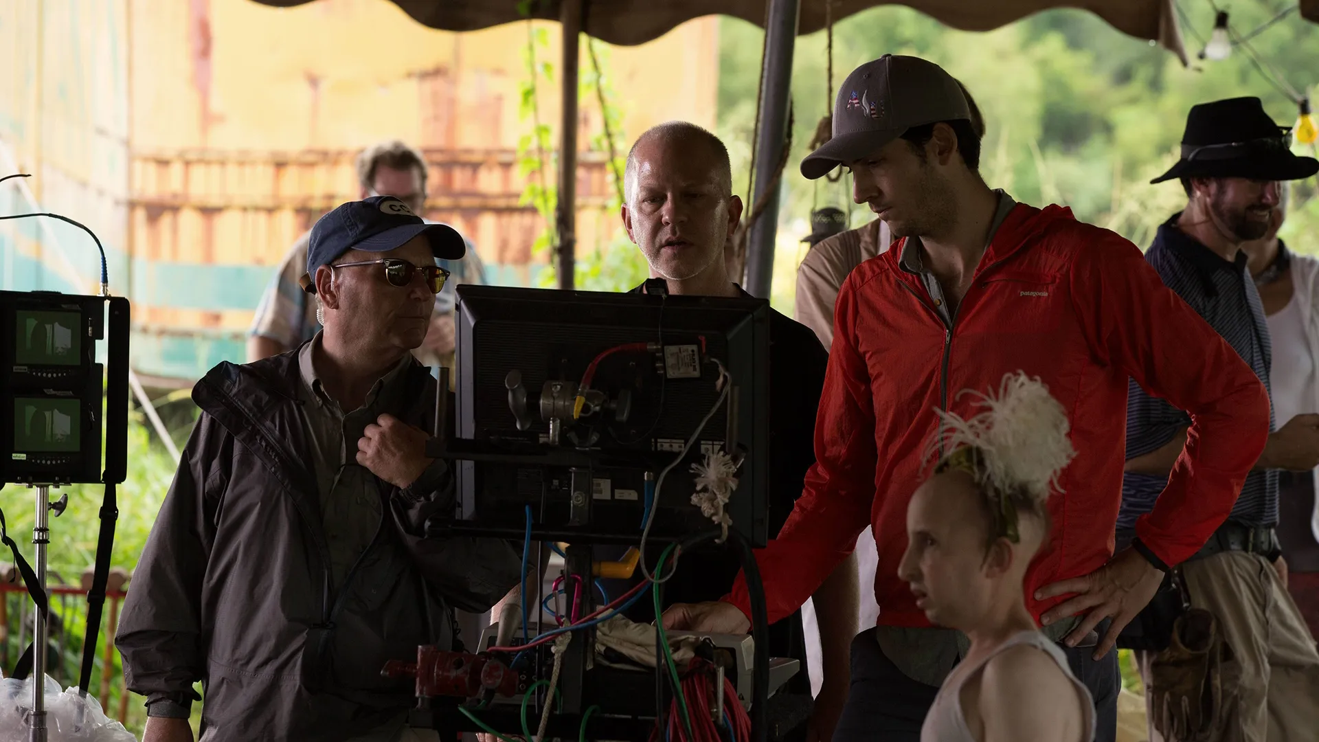 Show creator Ryan Murphy stands behind a camera monitor alongside a production crew and actor Meep (Ben Woolf) on an outdoor film set, capturing a candid, behind-the-scenes moment during the production of American Horror Story: Freak Show.