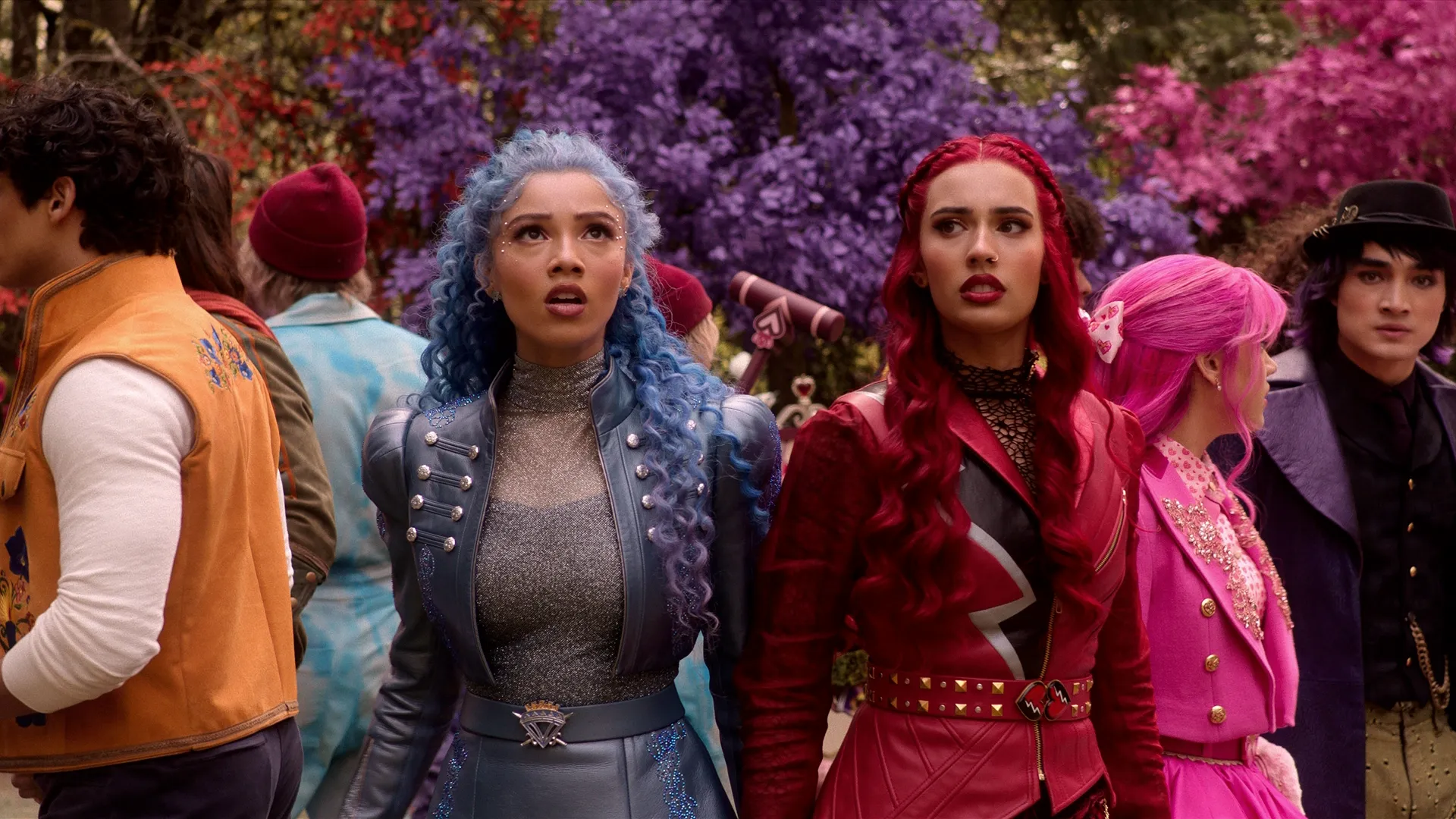 Chloe Charming (Malia Baker) and Red (Kylie Cantrall) look upward with wide eyes and stunned expressions as they stand outside surrounded by an assortment of colorful fairytale characters in a vibrant, purple and pink wonderland setting.