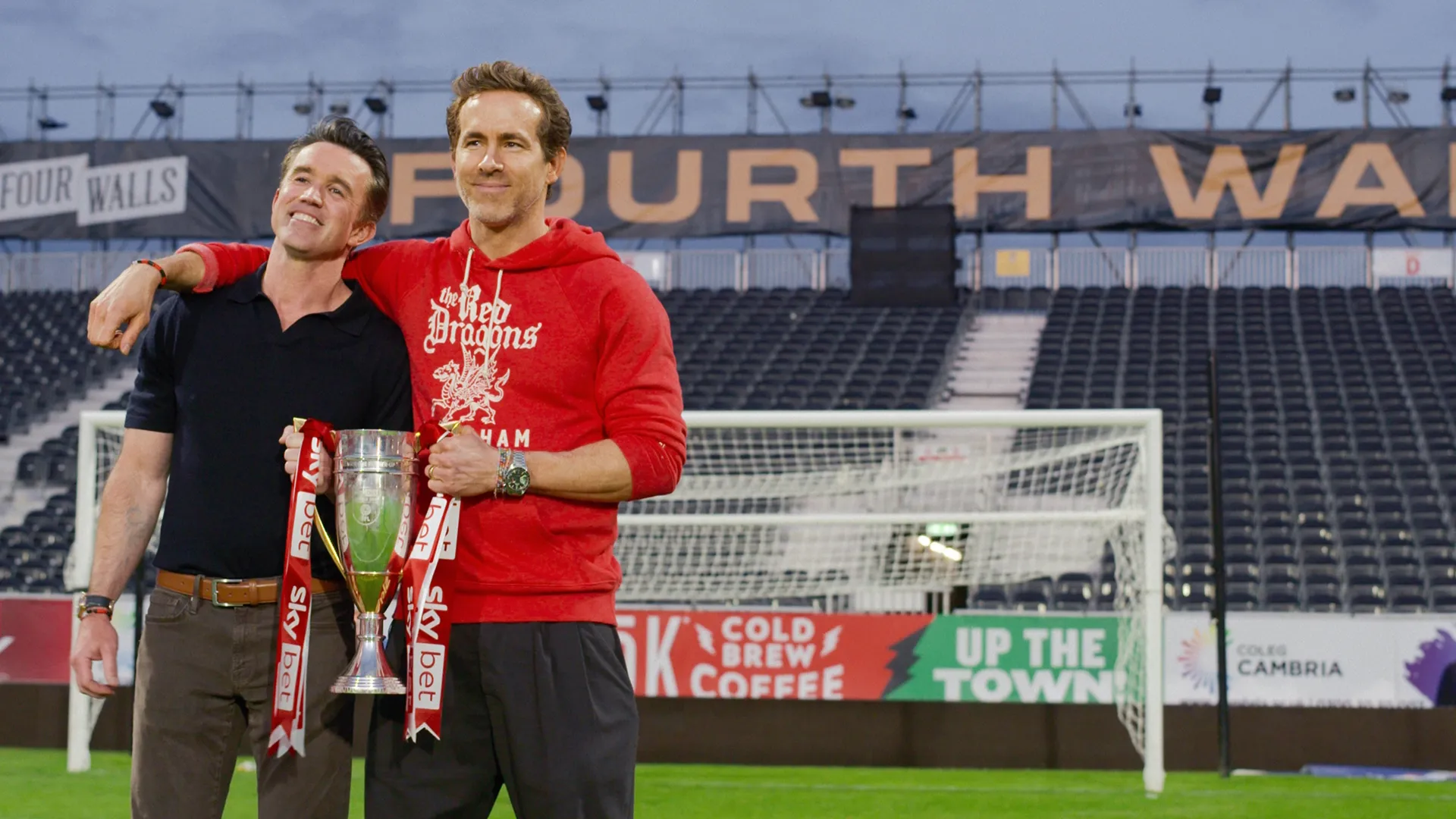 A celebratory on-field scene from the sports docuseries Welcome to Wrexham showing Rob McElhenney and Ryan Reynolds smiling with their arms around each other while holding up a silver Sky Bet promotion trophy inside the Wrexham stadium.