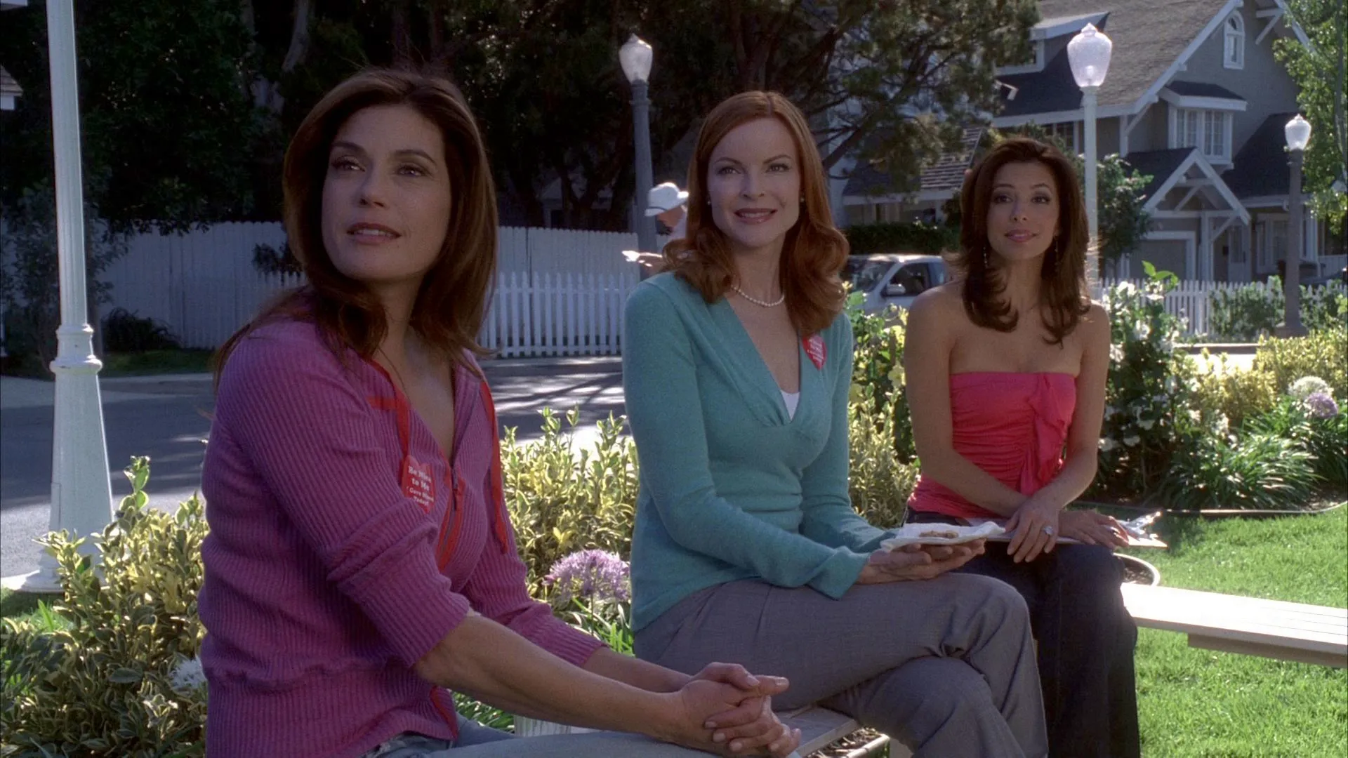 A scene from "Desperate Housewives" showing Susan, Bree, and Gabrielle sitting outdoors on a suburban lawn, smiling as they look off-camera.