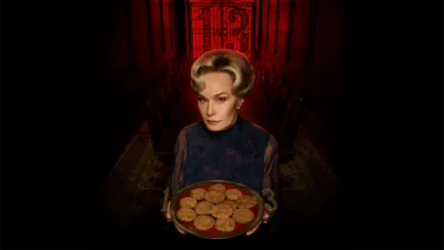 Actress Jessica Lange stands inside a dark, red-lit corridor holding a tray of cookies, with the number 13 visible in the background gate and on the tray handles.