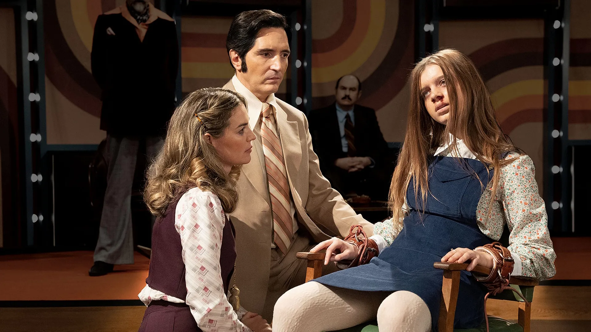 A man and a woman in 1970s clothing, kneeling by a teen girl who is tied to a chair