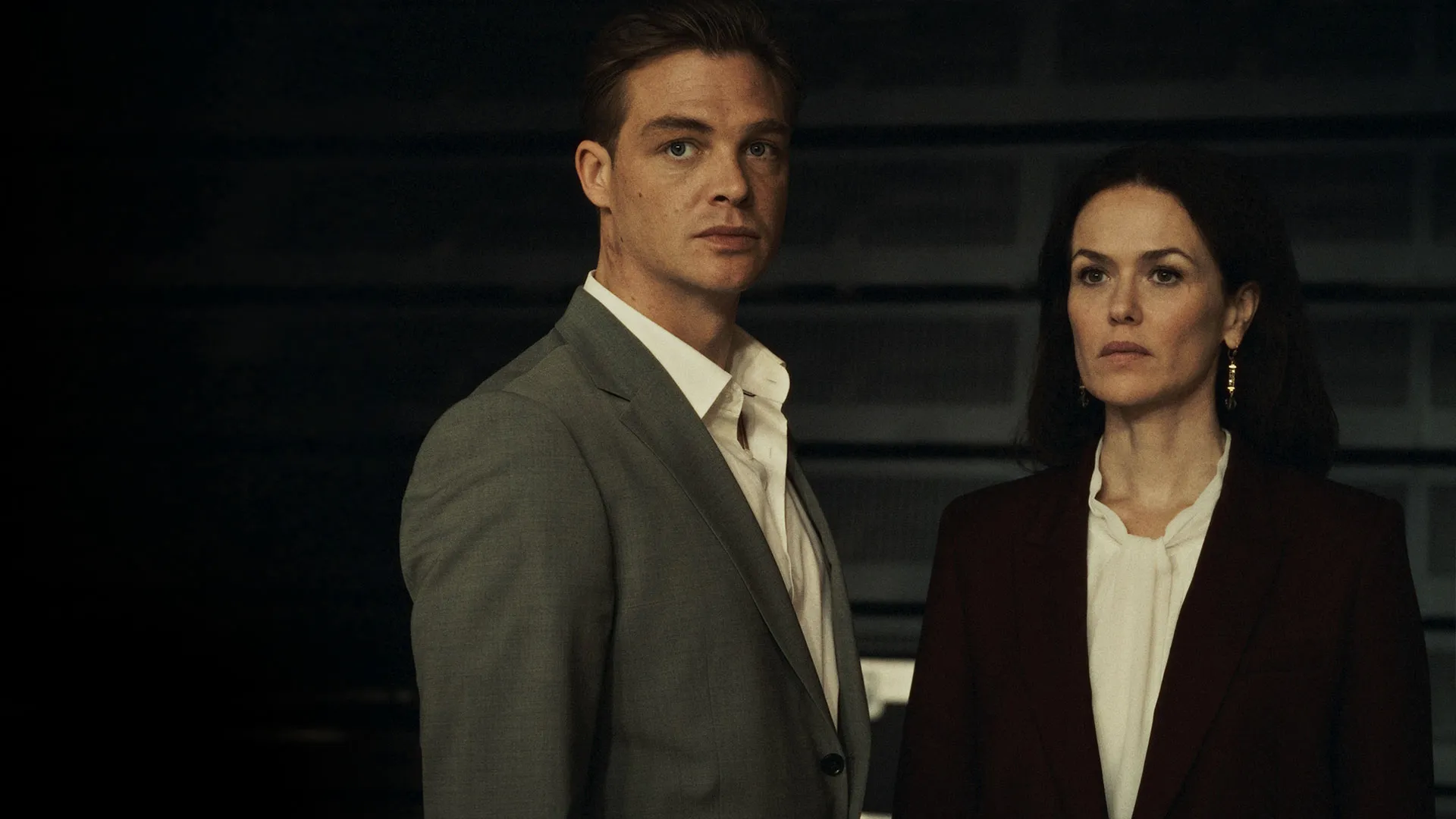 Two people in formal clothing stand side by side in a dark interior.