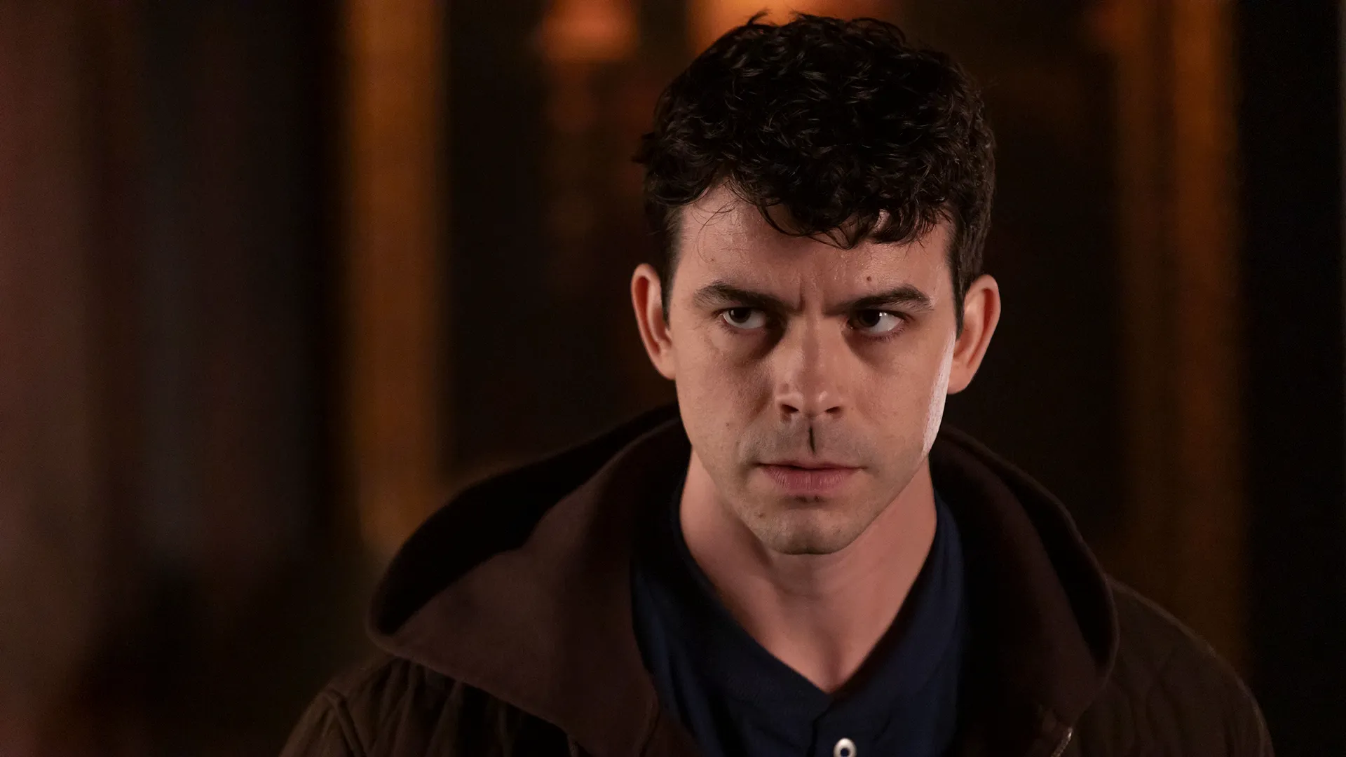 A young man with short curly dark hair and an intense expression looks slightly to the side, wearing a dark blue buttoned shirt under a brown hooded jacket.