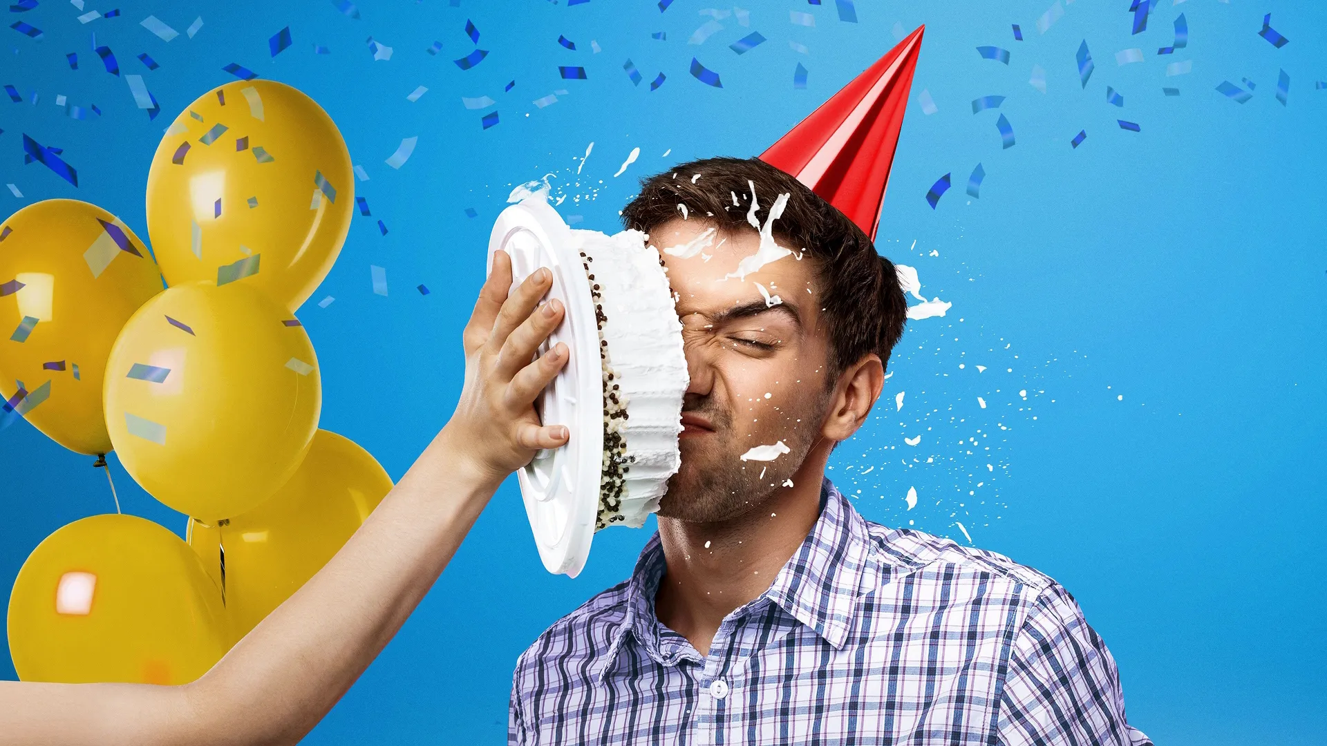 A man has a cake thrown in his face while wearing a red party hat. Yellow balloons and confetti float in the background.