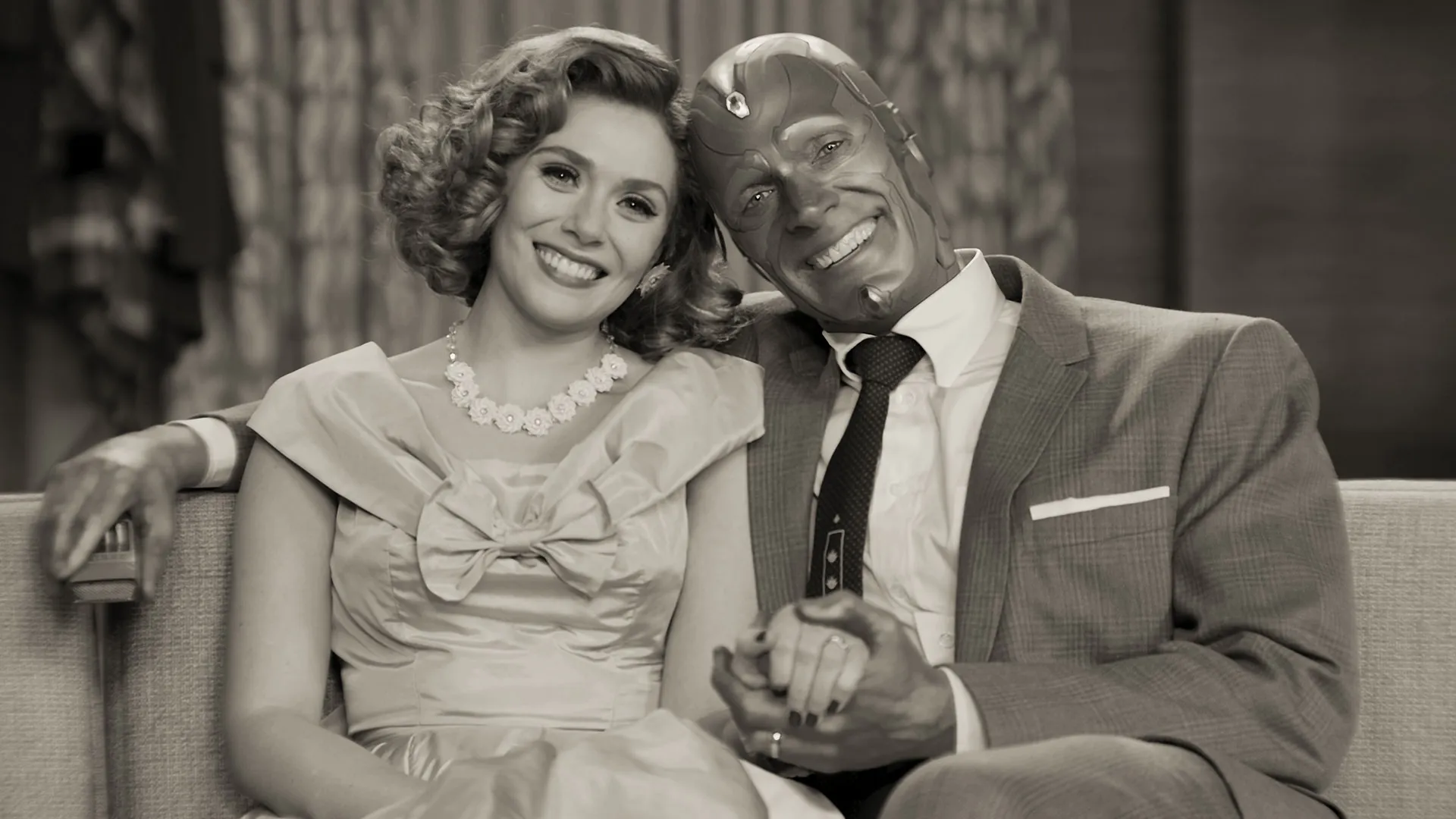 Wanda Maximoff and Vision, played by Elizabeth Olsen and Paul Bettany, smile warmly while sitting closely together on a couch in a black-and-white, 1950s sitcom-style scene from Marvel Studios' WandaVision.