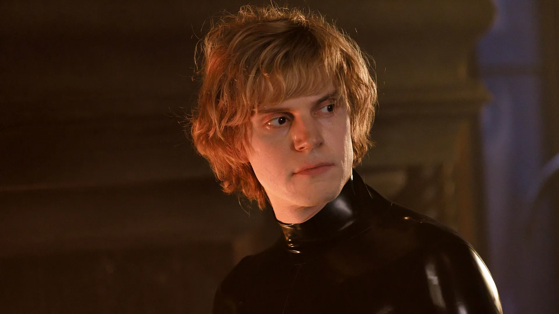 A young man with shaggy blonde hair looks off to the side with an intense expression, wearing a tight, high-collared black latex bodysuit.