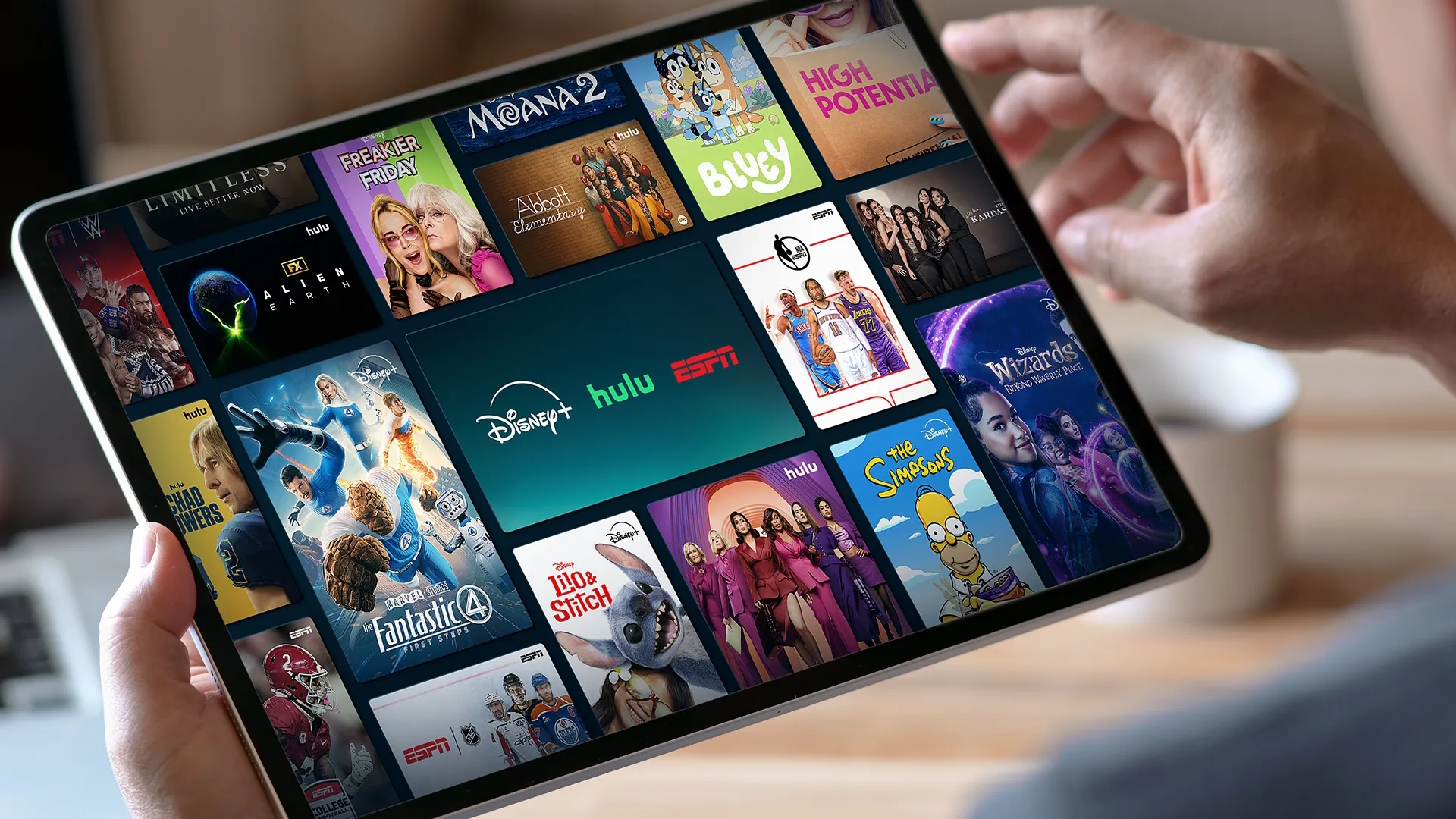 Closeup of a tablet device in someone's hands, the screen features the Disney+, Hulu, ESPN bundle logo in the center with key art from select titles that are available with the bundle around it.