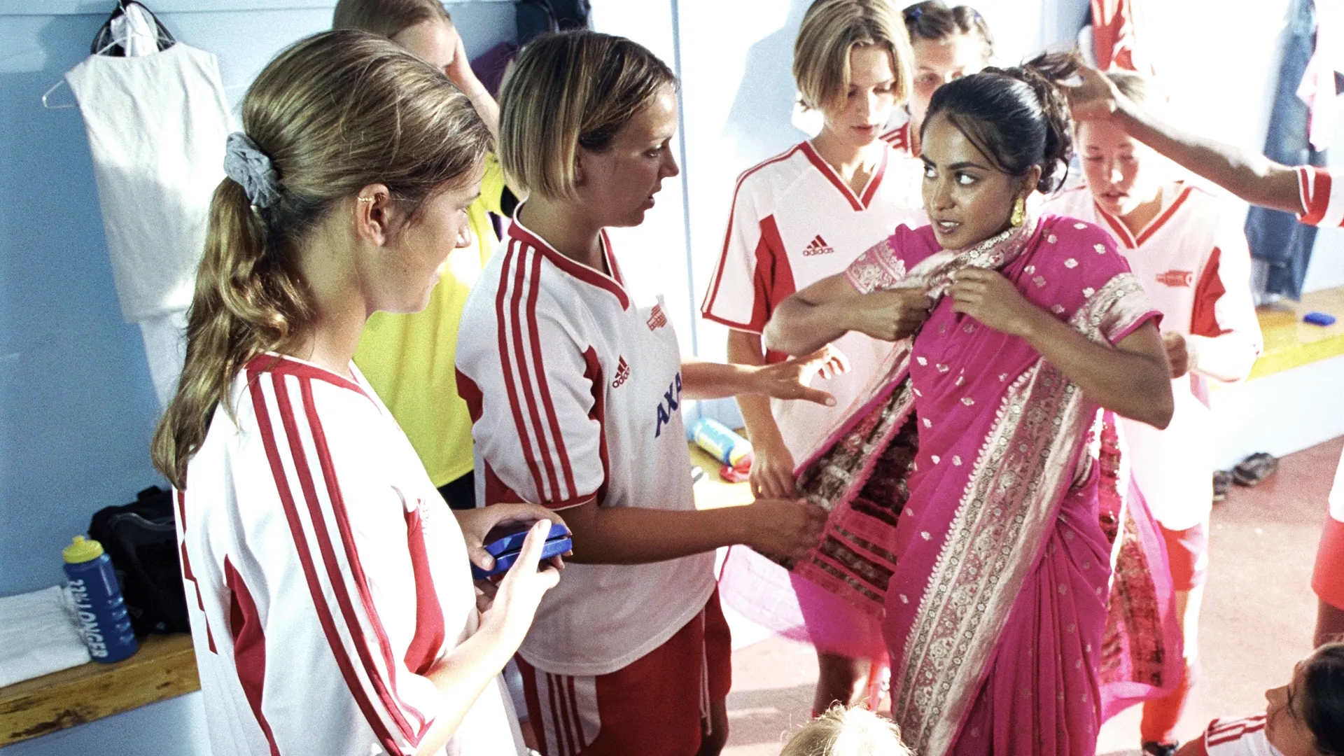 This scene depicts a moment from the 2002 film Bend It Like Beckham, featuring the character Jess Bhamra dressed in a traditional pink saree while her teammates look on in a locker room setting.