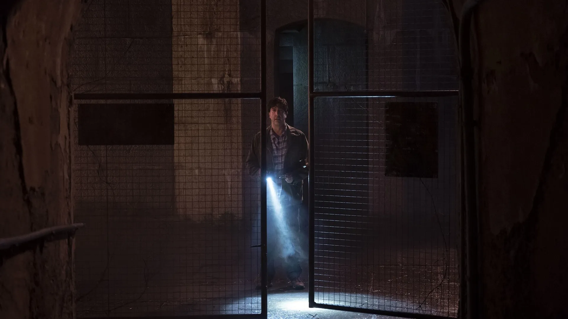 Davis Schwimmer's character investigates an open chain-link fence with a flashlight in the dark.