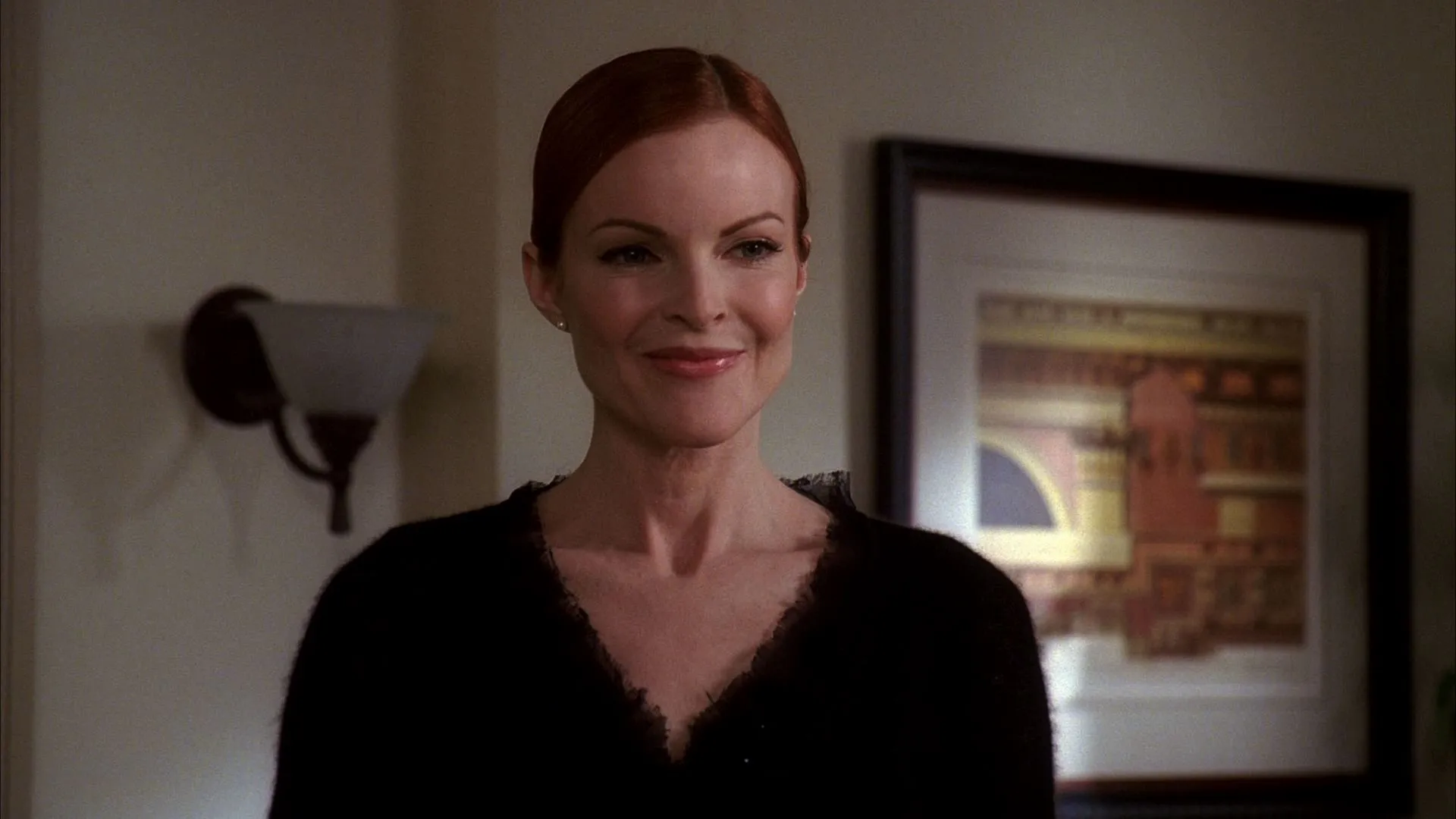 A scene from "Desperate Housewives" featuring Bree looking directly forward with a soft, polite smile, wearing a black v-neck top with her red hair neatly pulled back, set against a softly lit indoor background with a framed picture.