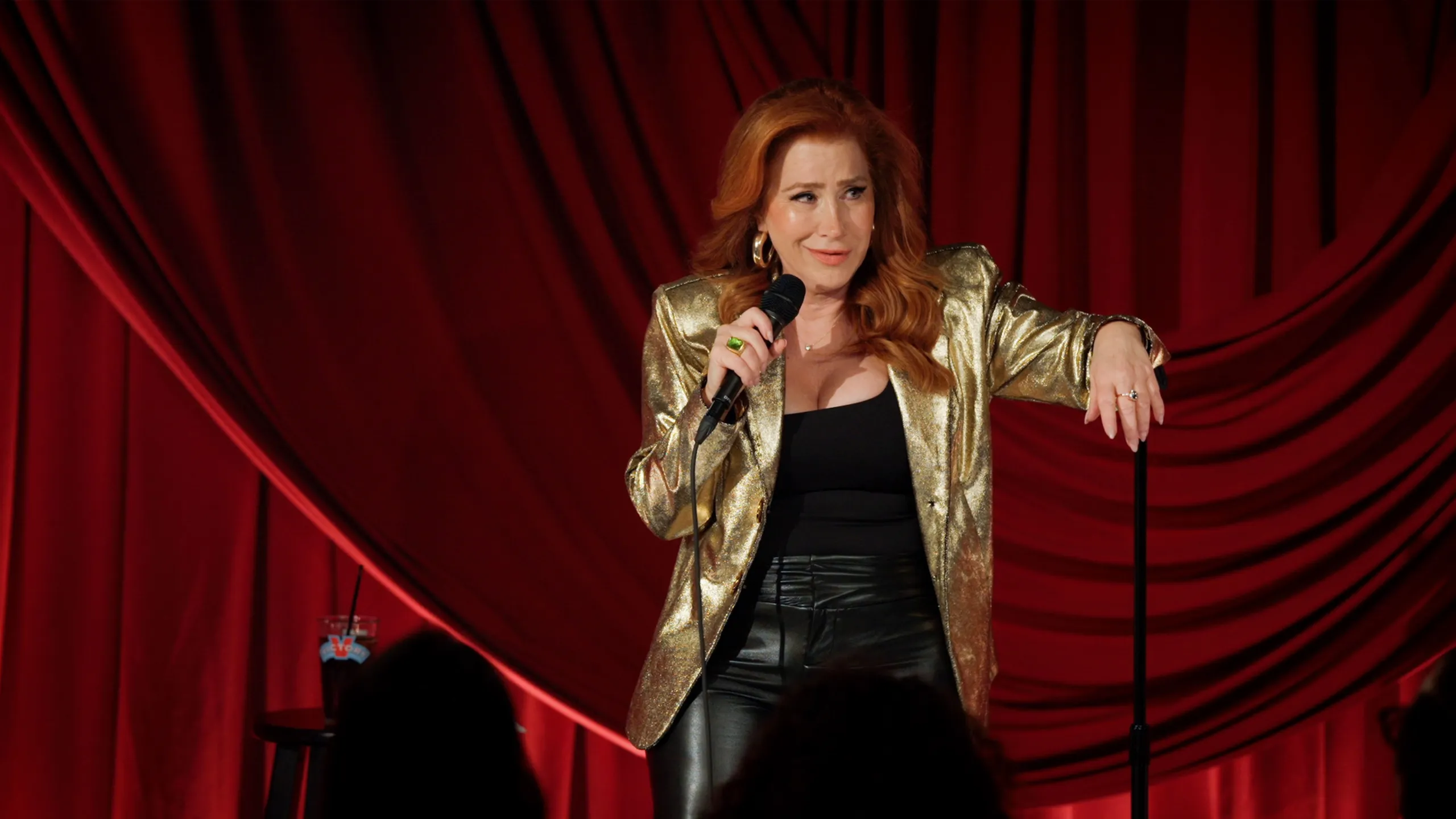 Lisa Ann Walter performs on stage holding a microphone in front of red curtains in Lisa Ann Walter: It Was An Accident on Disney+.