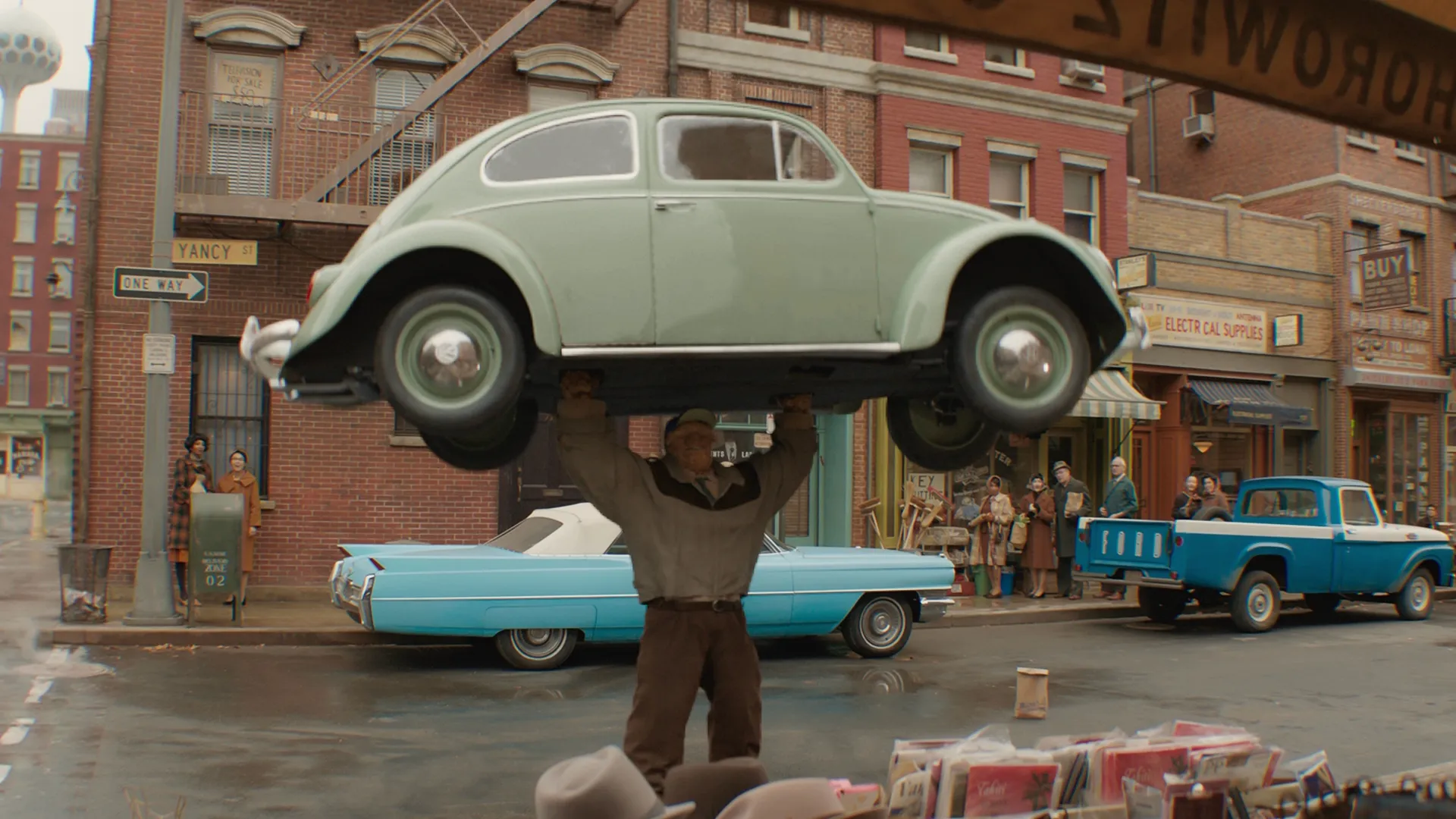 The Thing stands in the street, holding a 60s-looking car above his head as a small group of people look on