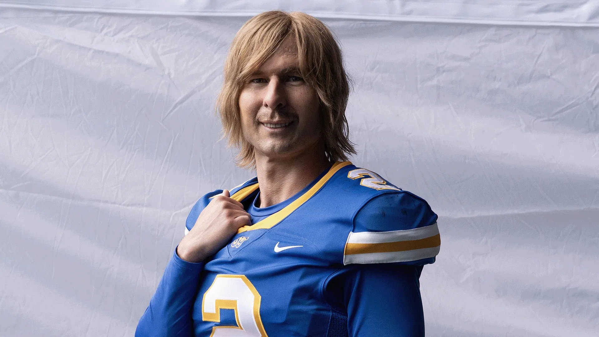 Actor Glen Powell wearing a blonde wig and a blue number 2 "South Georgia Catfish" football jersey in a promotional poster for the Hulu sports comedy series "Chad Powers