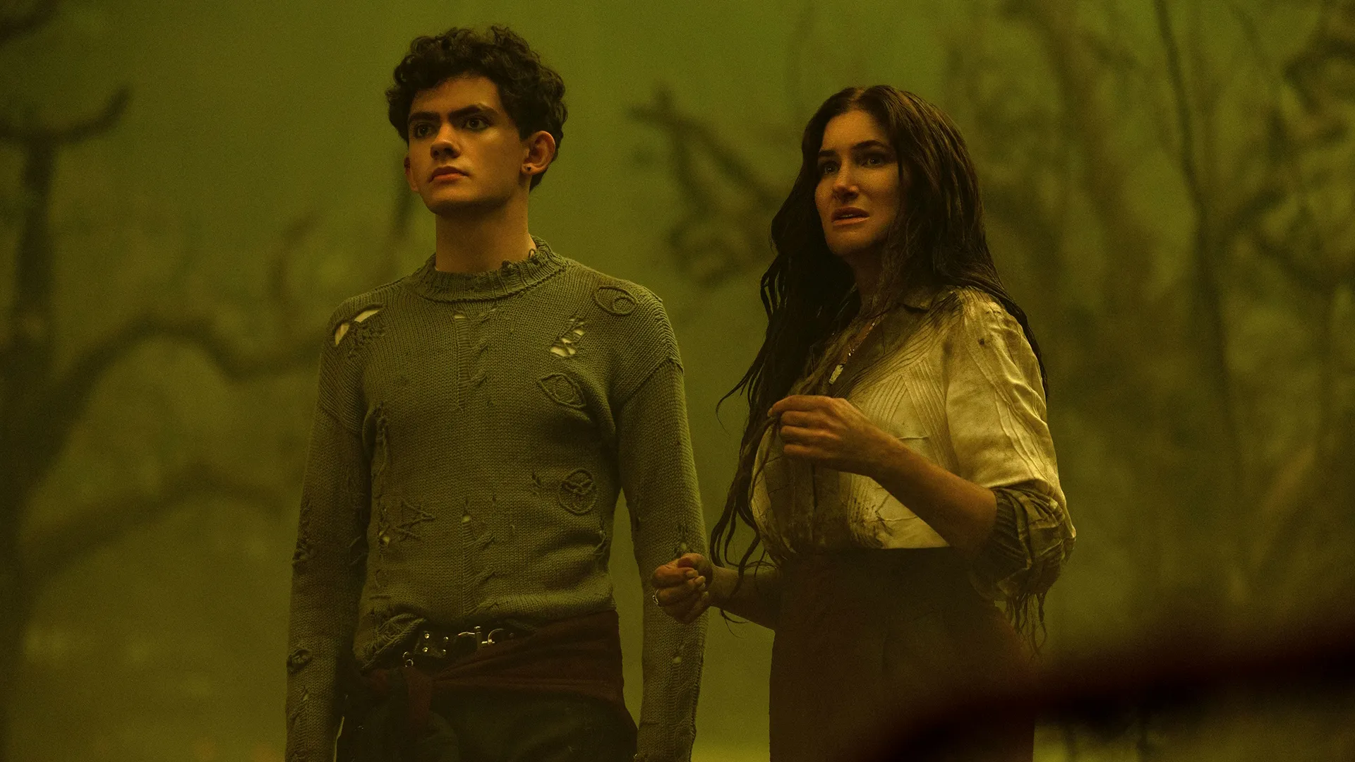Teen / Billy Maximoff (Joe Locke) and Agatha Harkness (Kathryn Hahn) stand close together, looking off-camera with concerned and alert expressions against a hazy, sickly green-lit forest backdrop.