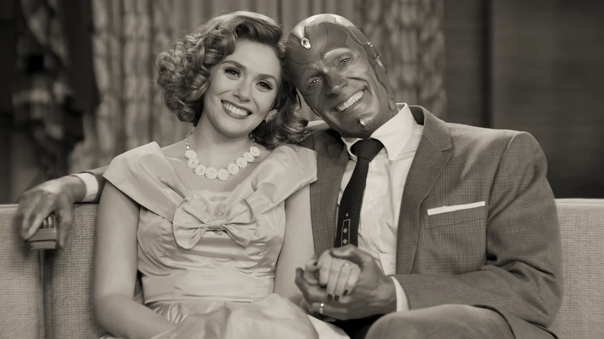 Wanda Maximoff and Vision, played by Elizabeth Olsen and Paul Bettany, smile warmly while sitting closely together on a couch in a black-and-white, 1950s sitcom-style scene from Marvel Studios' WandaVision.
