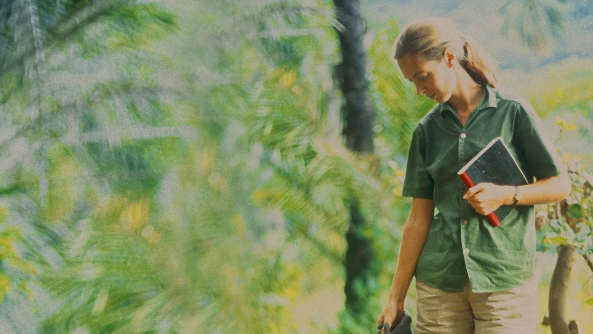 A young Jane Goodall walks through a tropical forest holding a notebook