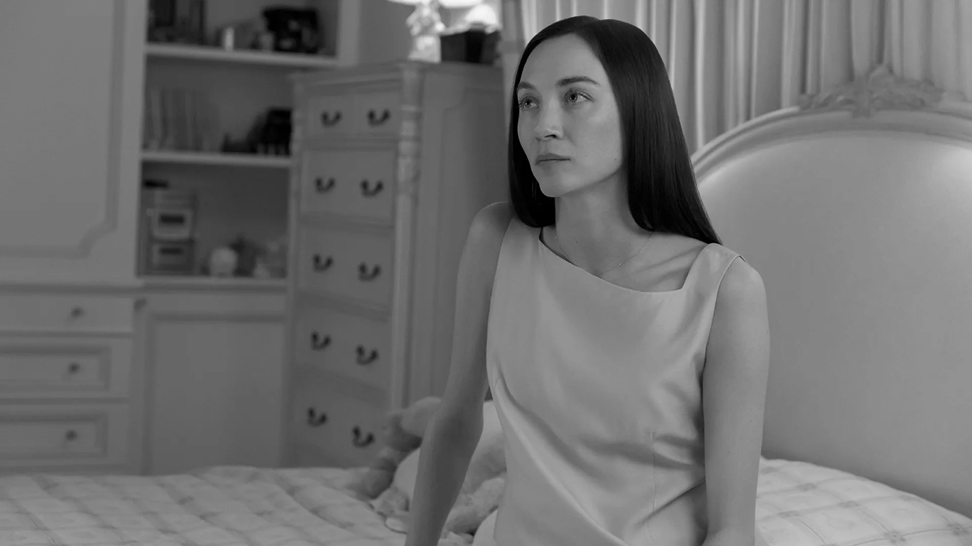 A brunette Carolyn Bessette (Sarah Pidgeon) sitting on a bed in a black and white set photo from FX's Love Story.