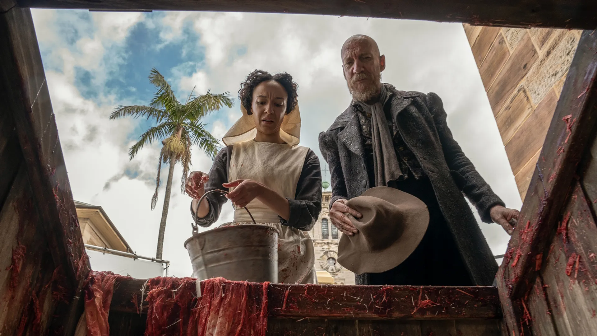 Marianne (Andrea Demetriades) and Fagin (David Thewlis) look down into a the opening of a blood-stained wooden pit