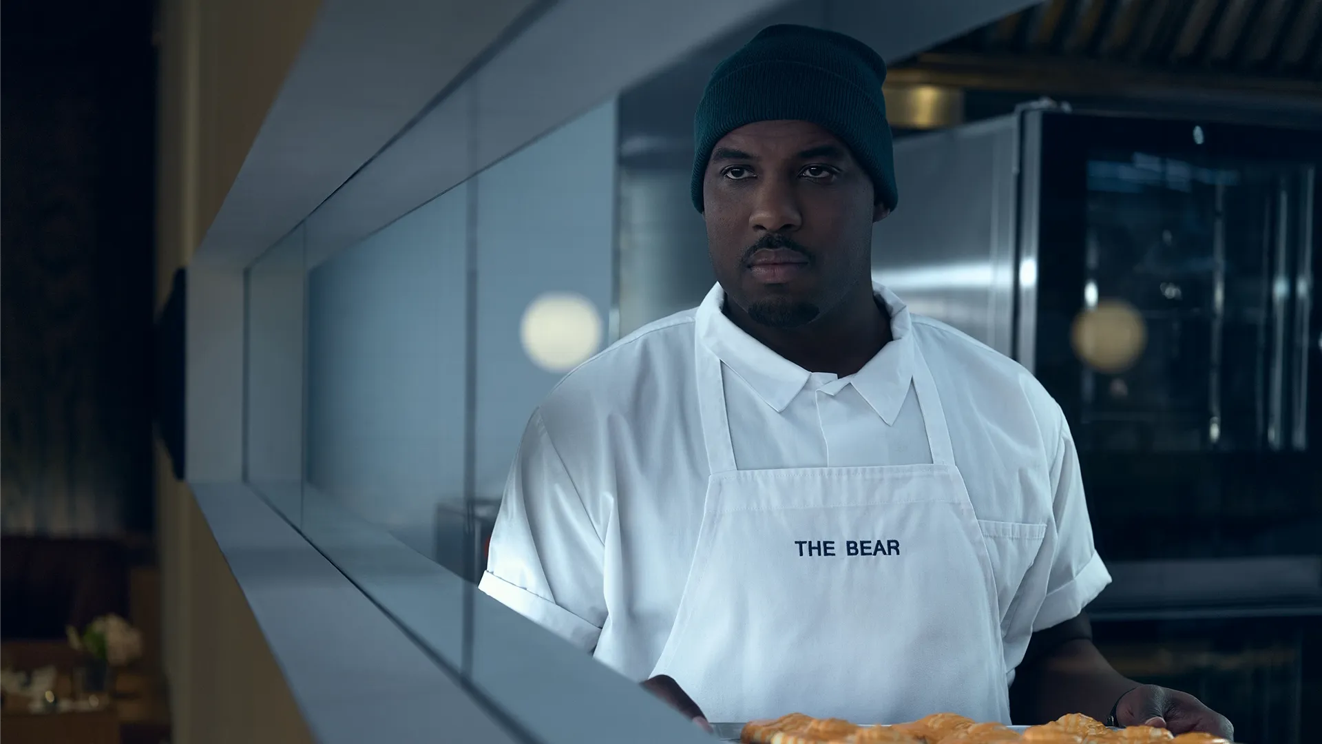 Plano médio de Marcus Brooks (Lionel Boyce) sério, vestindo um gorro azul e um avental de chef branco bordado com "O URSO", olhando para fora do plano da câmera enquanto segura uma bandeja de panificados em uma cozinha profissional.