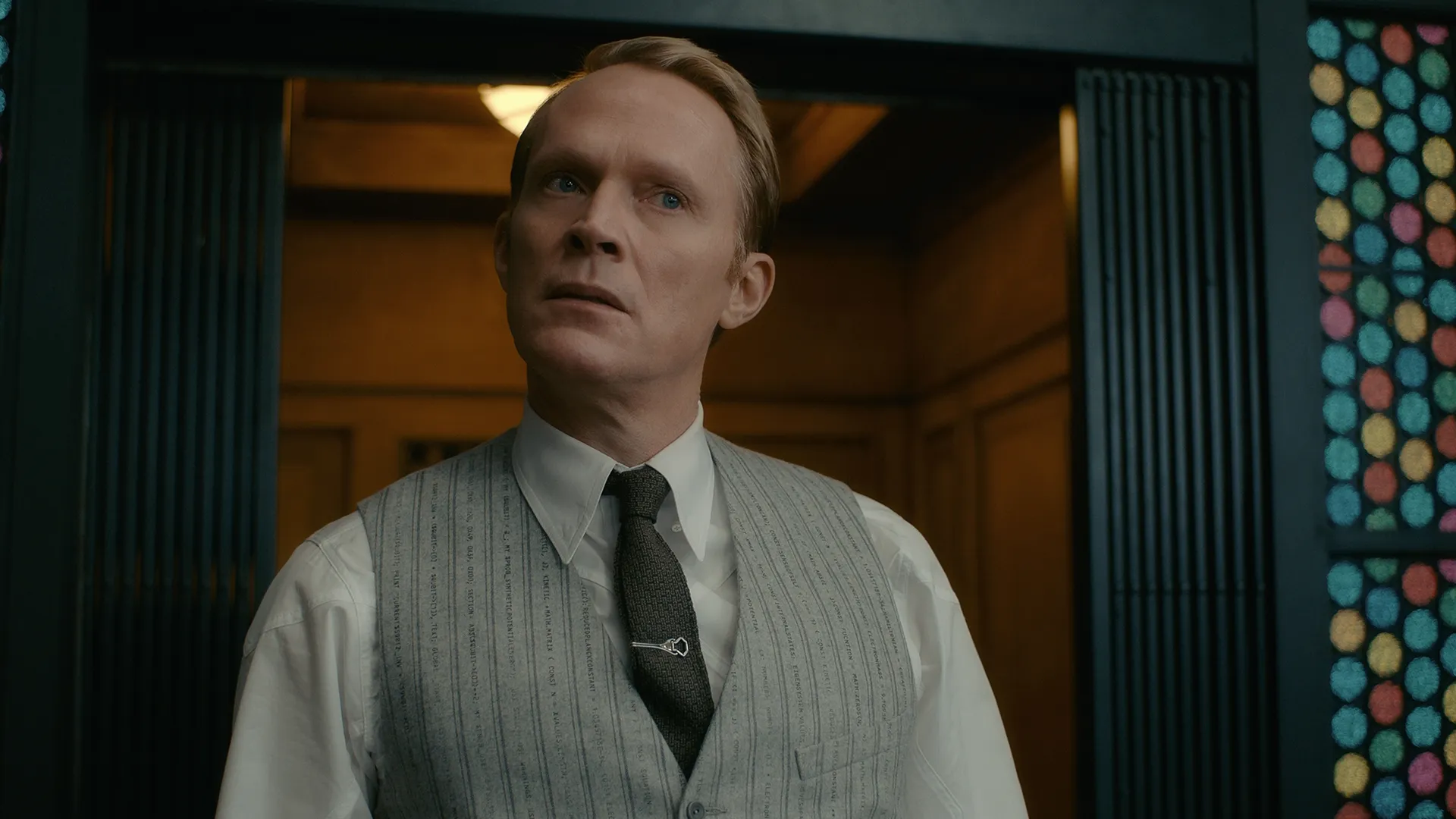 Vision, played by Paul Bettany, stands in a doorway wearing a light gray pinstriped vest inscribed with quantum physics equations, a white button-down shirt, and a dark tie secured by a tie clip in Marvel's VisionQuest.