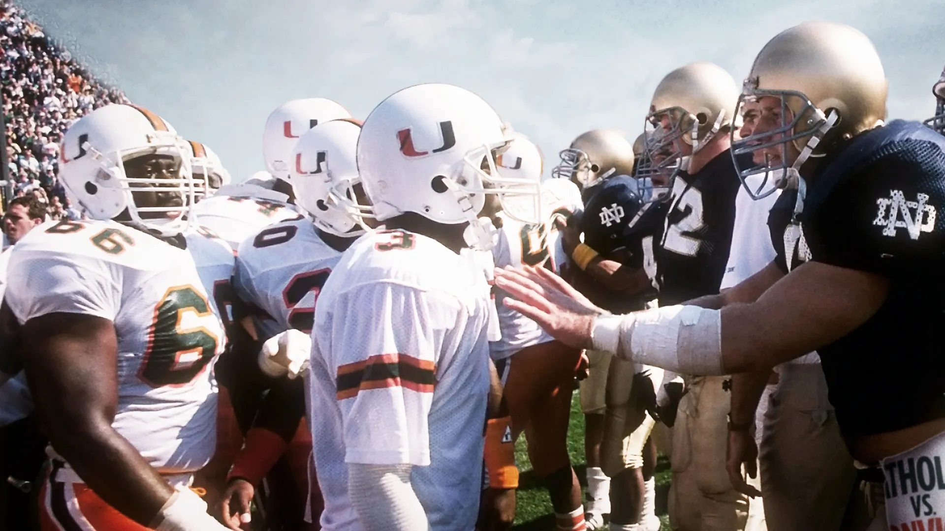 An American football player from the Miami Hurricanes and a player from the Notre Dame Fighting Irish stand face-to-face on the field amidst their teammates.