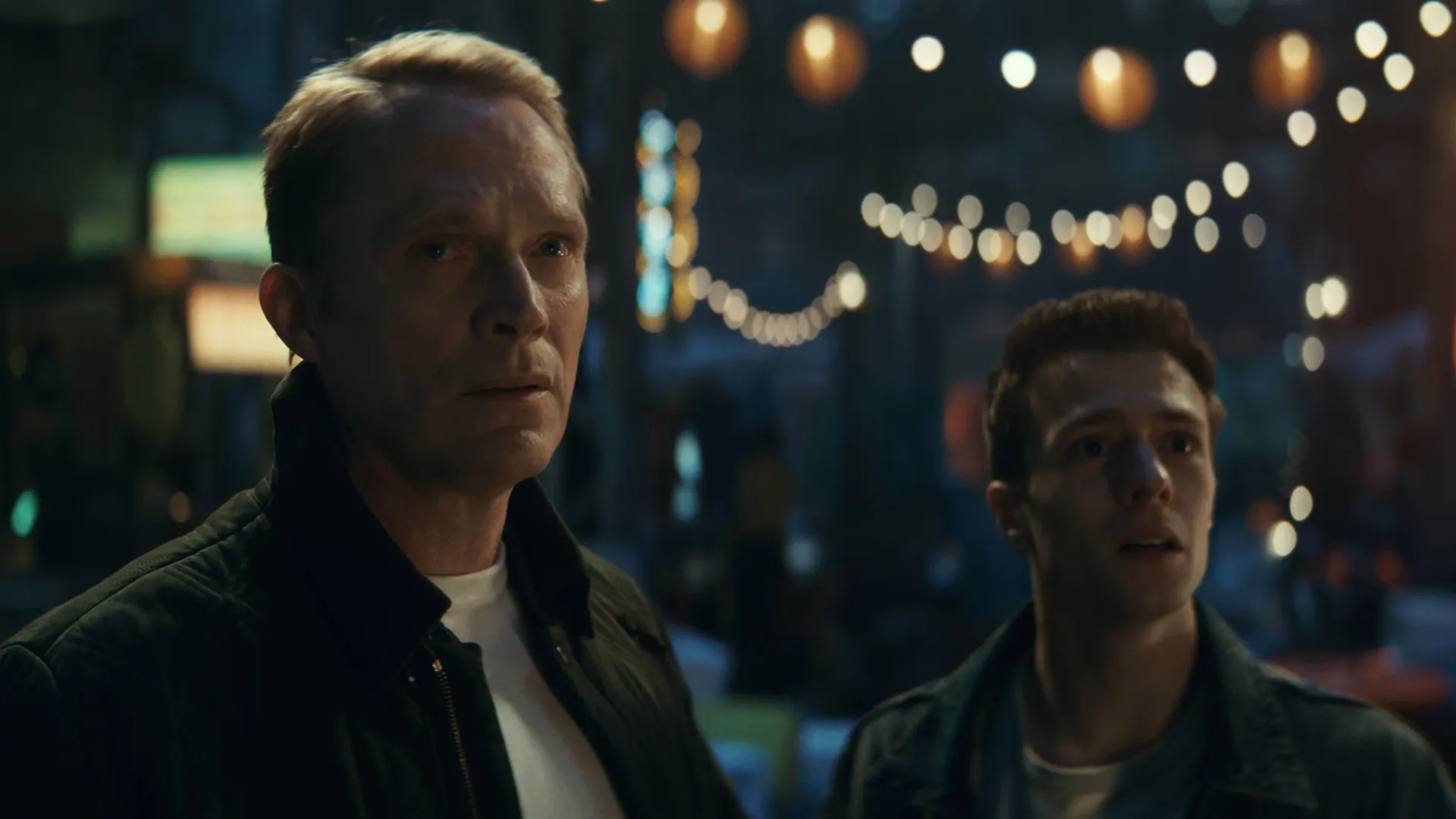 Vision and Tommy Maximoff stand side-by-side with expressions of concern and surprise against a blurred backdrop of string lights in Marvel's VisionQuest.