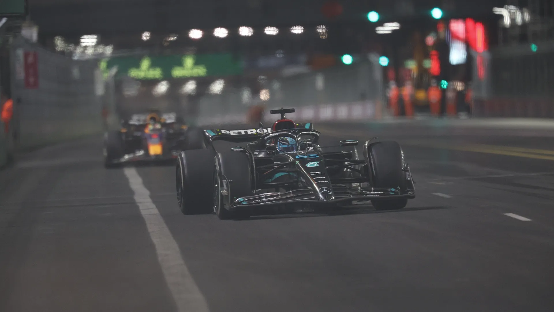 The black livery Mercedes of George Russell (No. 63) is pursued by the Red Bull of Max Verstappen (No. 1) during a night race at a street circuit.