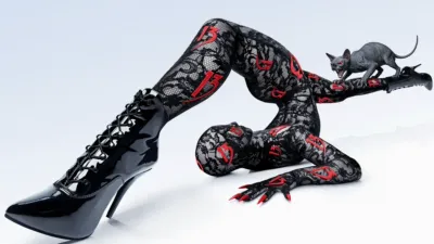 A contortionist posing on a white background wears a black lace bodysuit patterned with red number 13s, black patent leather stiletto boots, and long red fingernails, while a small, hairless cat figure sits on one of their raised feet.