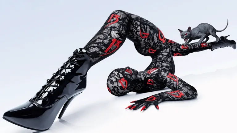 A contortionist posing on a white background wears a black lace bodysuit patterned with red number 13s, black patent leather stiletto boots, and long red fingernails, while a small, hairless cat figure sits on one of their raised feet.