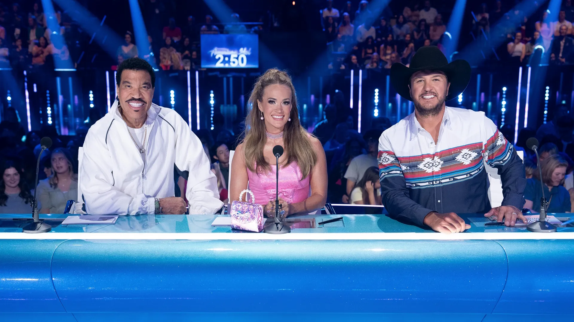 American Idol judges Lionel Richie, Carrie Underwood, and Luke Bryan sit behind the judges' desk, smiling at the camera