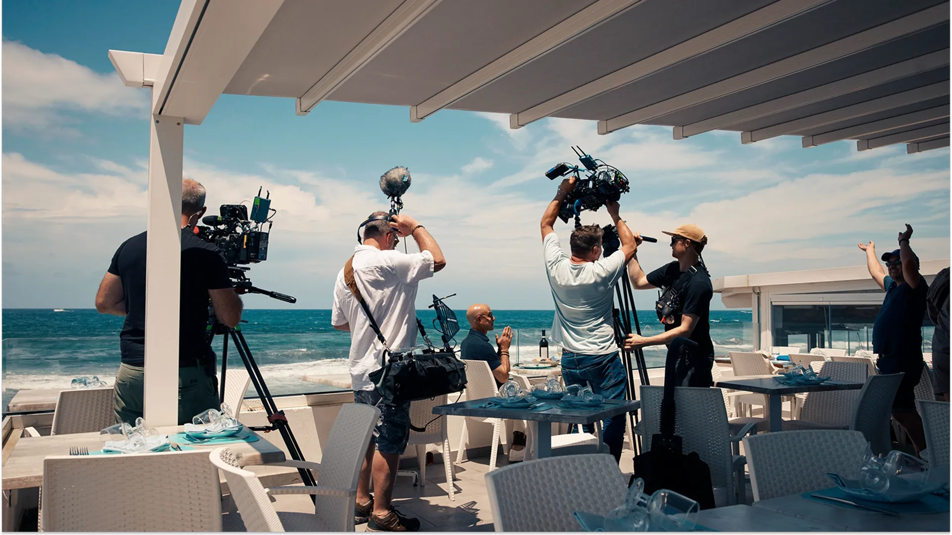 A camera crew films Stanley Tucci sat at a dining table in the outside area of a restaurant by the sea