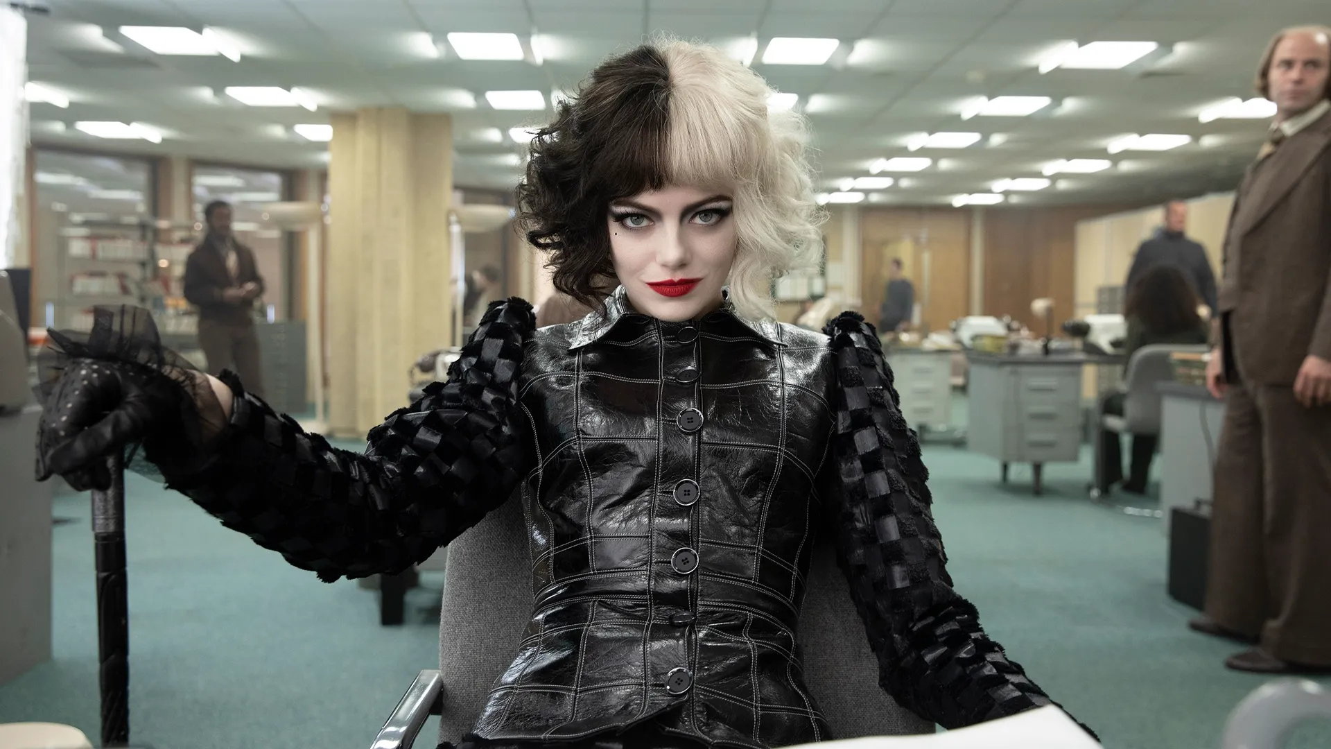 A young Cruella De Vil (Emma Stone) sits in an office with a devilish smirk.