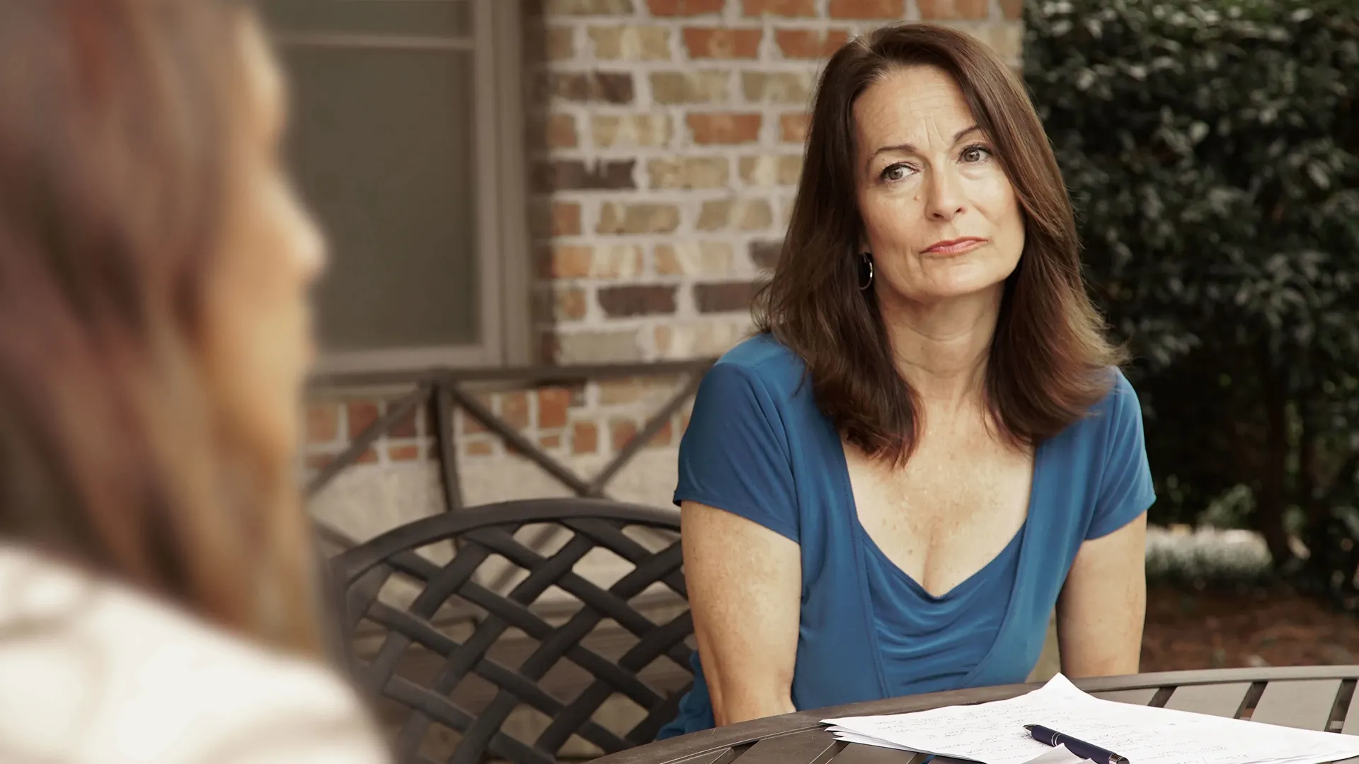 A woman with brown hair wearing a blue v-neck short-sleeve top while sitting outdoors near a brick wall looking at someone off-screen