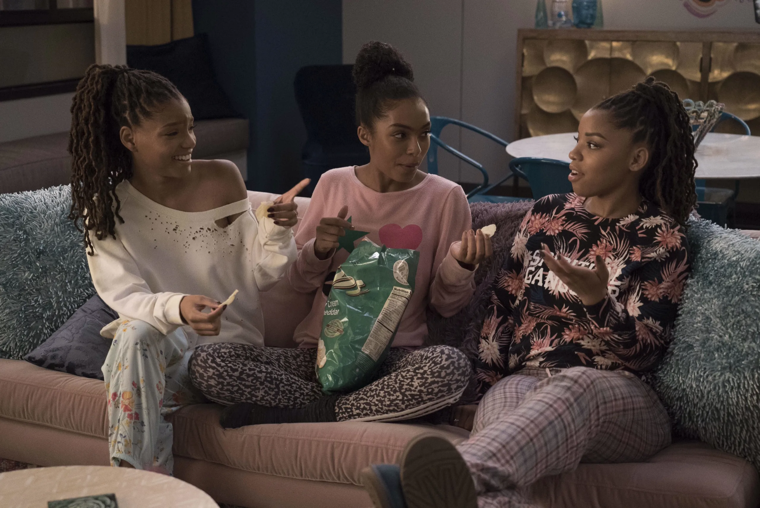 Three young women played by Yara Shahidi, Chloe and Halle Bailey, sit on a couch having a discussion and eating chips