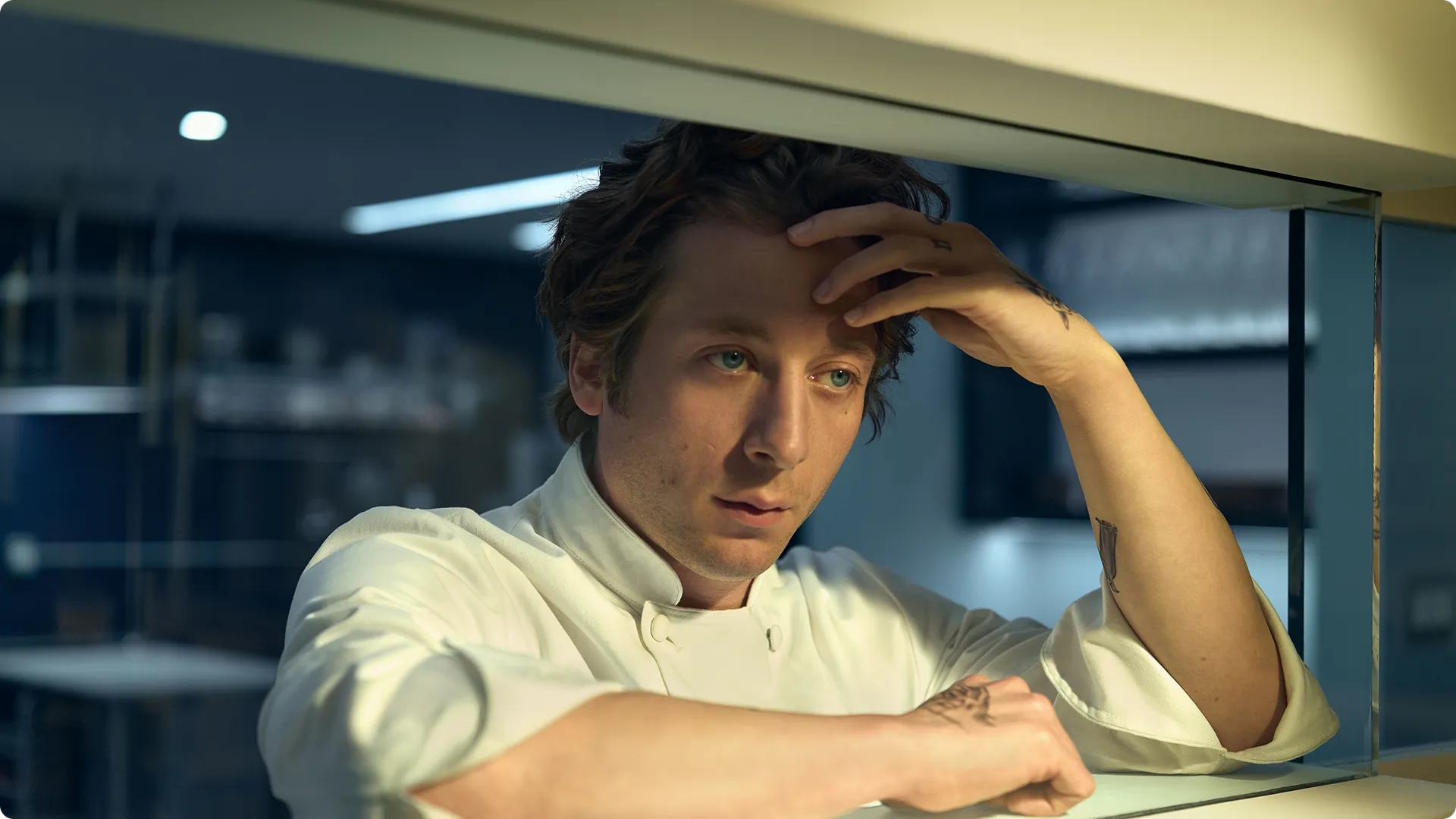 Close de Carmen "Carmy" Berzatto (Jeremy Allen White) pensativo, usando um jaleco branco de chef, apoiando os braços no balcão de uma cozinha de restaurante moderna enquanto repousa a testa na mão.