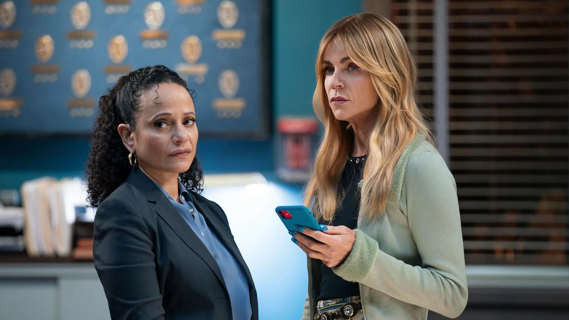 Morgan Gillory (Kaitlin Olson) and Selena Soto (Judy Reyes) are standing in a police station, looking seriously at something out of frame